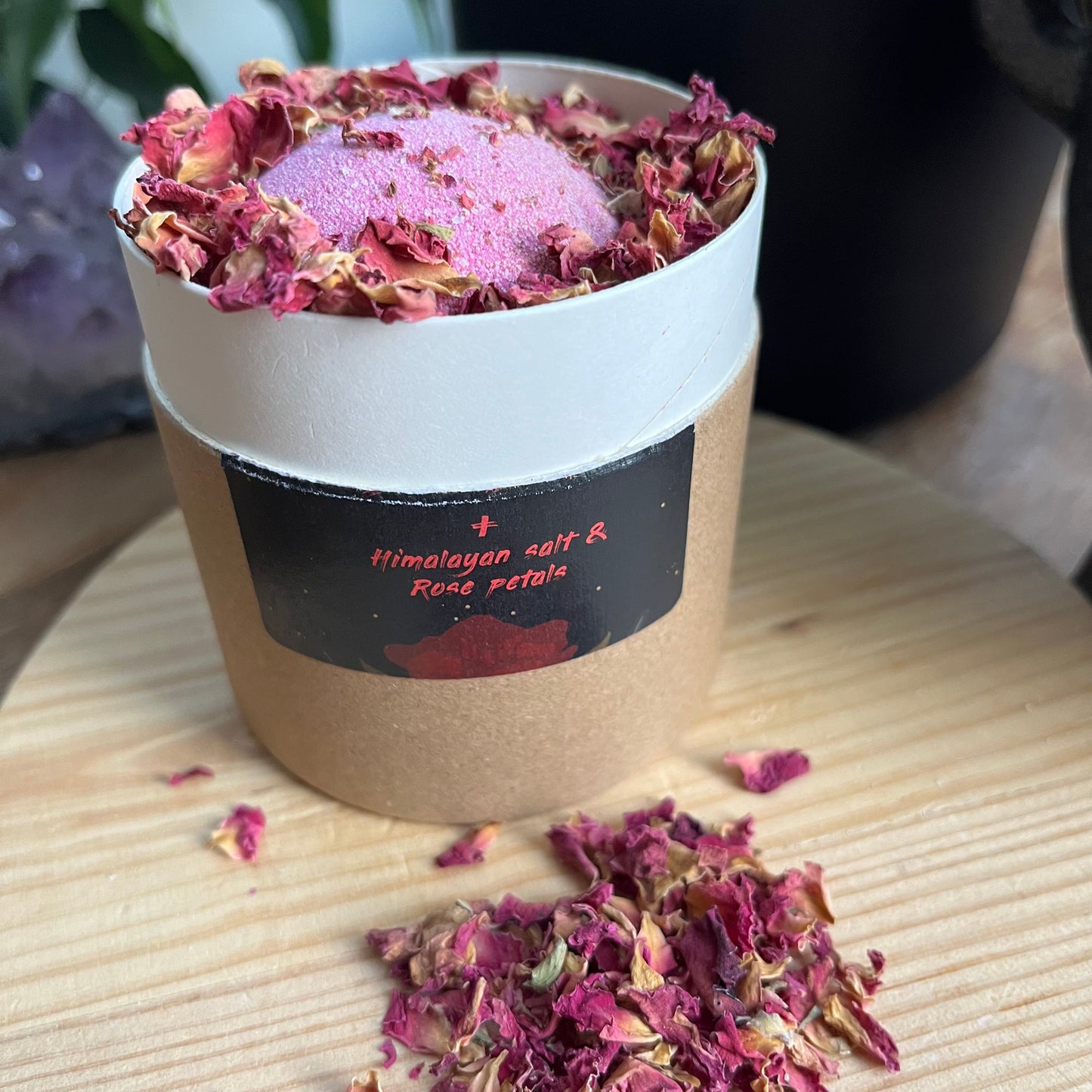 Circular cardboard presentation box containing a pink bath bomb and red flower petals