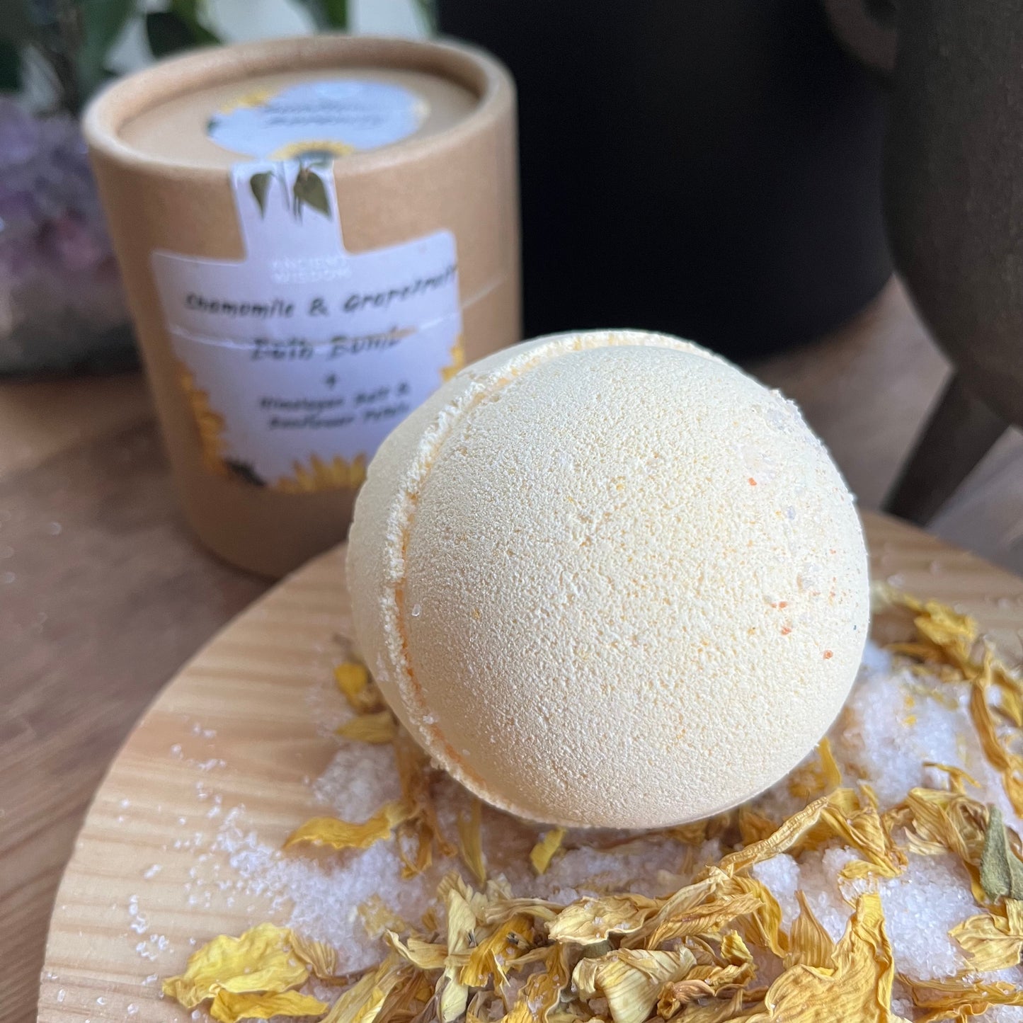 pale yellow bath bomb rested on top of yellow flower petals and white himalayan salt. in the background we can see the cardboard presentation box