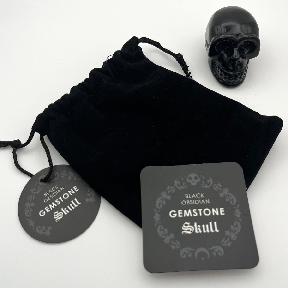 Black obsidian gemstone skull displayed with a black velvet drawstring pouch, a round black tag, and a square product card on a white background.
