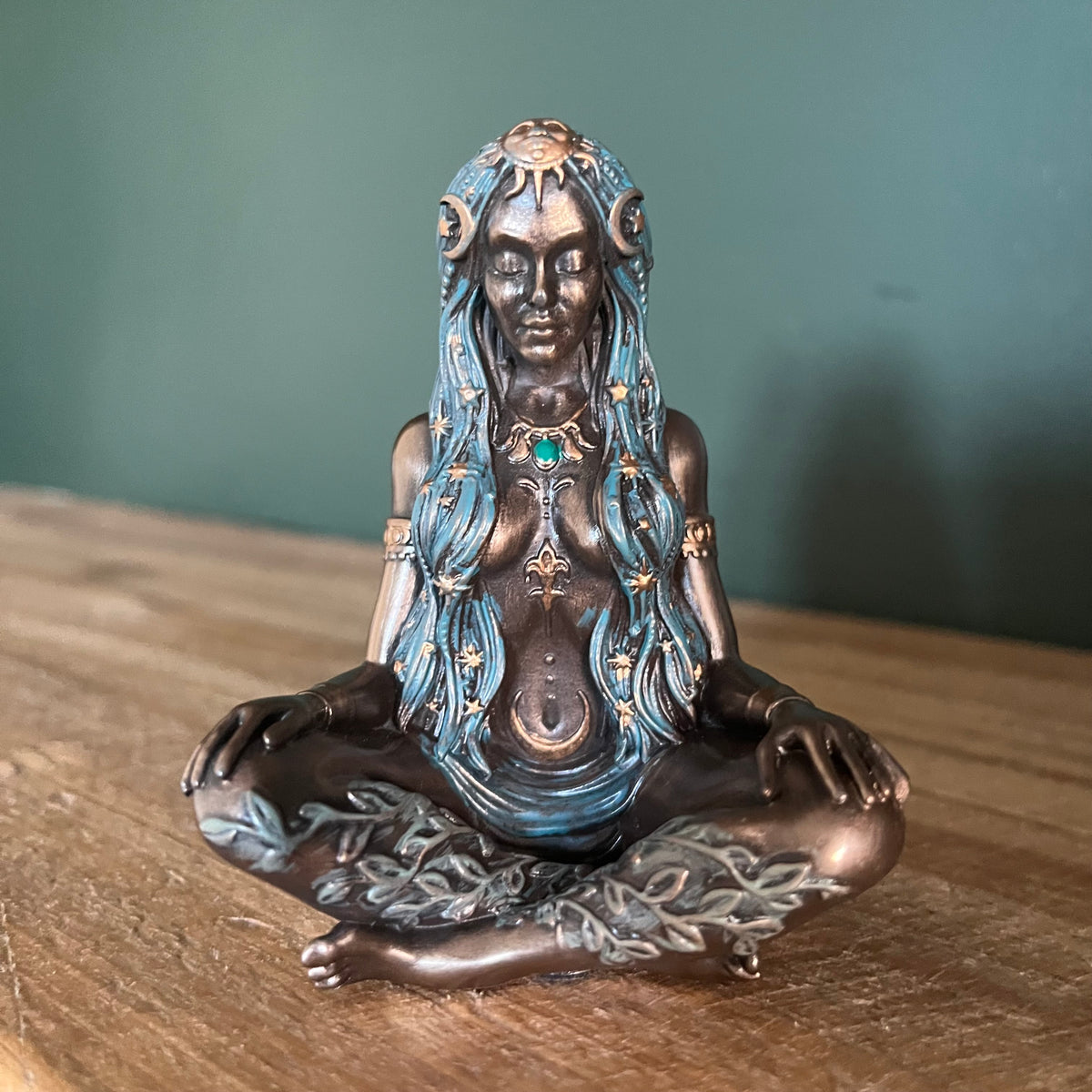 A bronze-finished female deity figurine in a meditative lotus pose, featuring blue patina highlights in her hair and chest, gold accents, and a green gemstone at her collarbone, sitting on a wooden surface against a dark green wall.