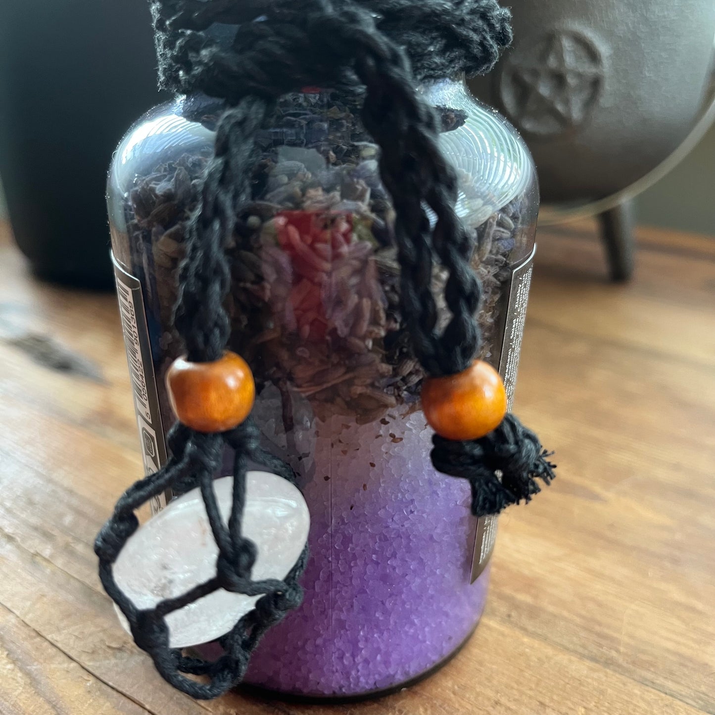 A glass jar candle with a black braided cord and wooden beads, featuring a white crystal pendant charm, set on a wooden surface.