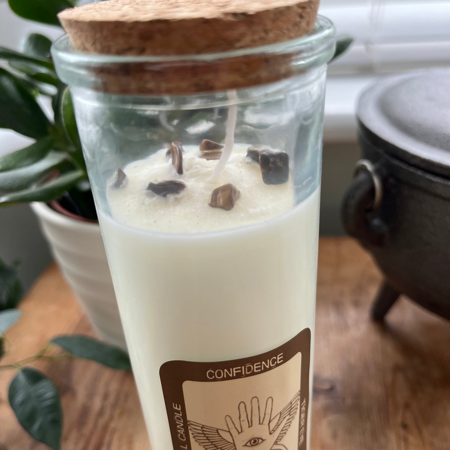 Tall glass spell candle with cork lid. The label on the candle features a winged hand with an eye in its palm and the words Confidence, Tigers eye and Cinnamon. The candle itself is white with Tigers eye gemstones embedded within it.