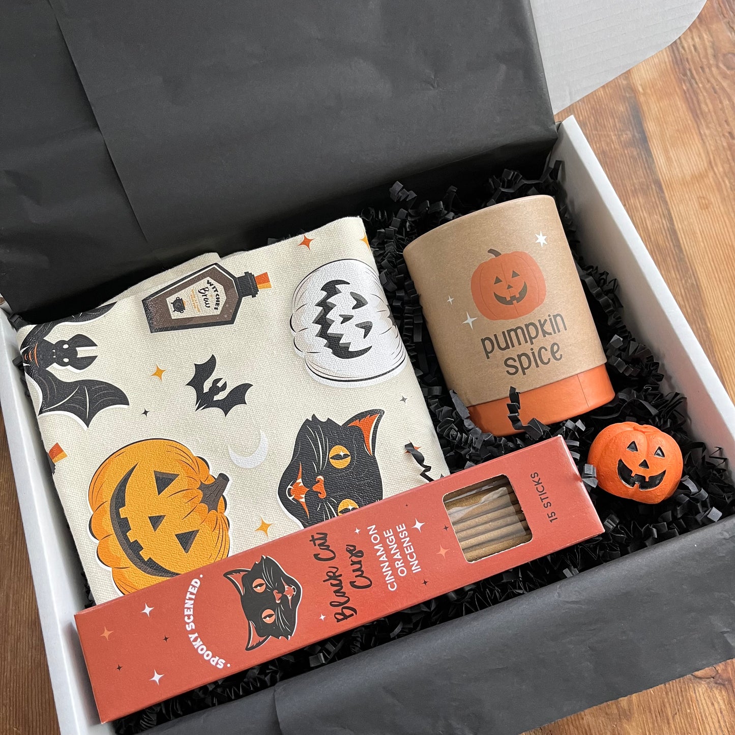 A curated Halloween-themed gift box containing a cream canvas tote bag with a black cat, jack-o'-lantern, and bat print; a boxed candle labeled 'pumpkin spice', box of 'Black Cat Cinnamon Orange' incense sticks; and a small orange jack-o'-lantern incense holder all arranged in black shredded paper.