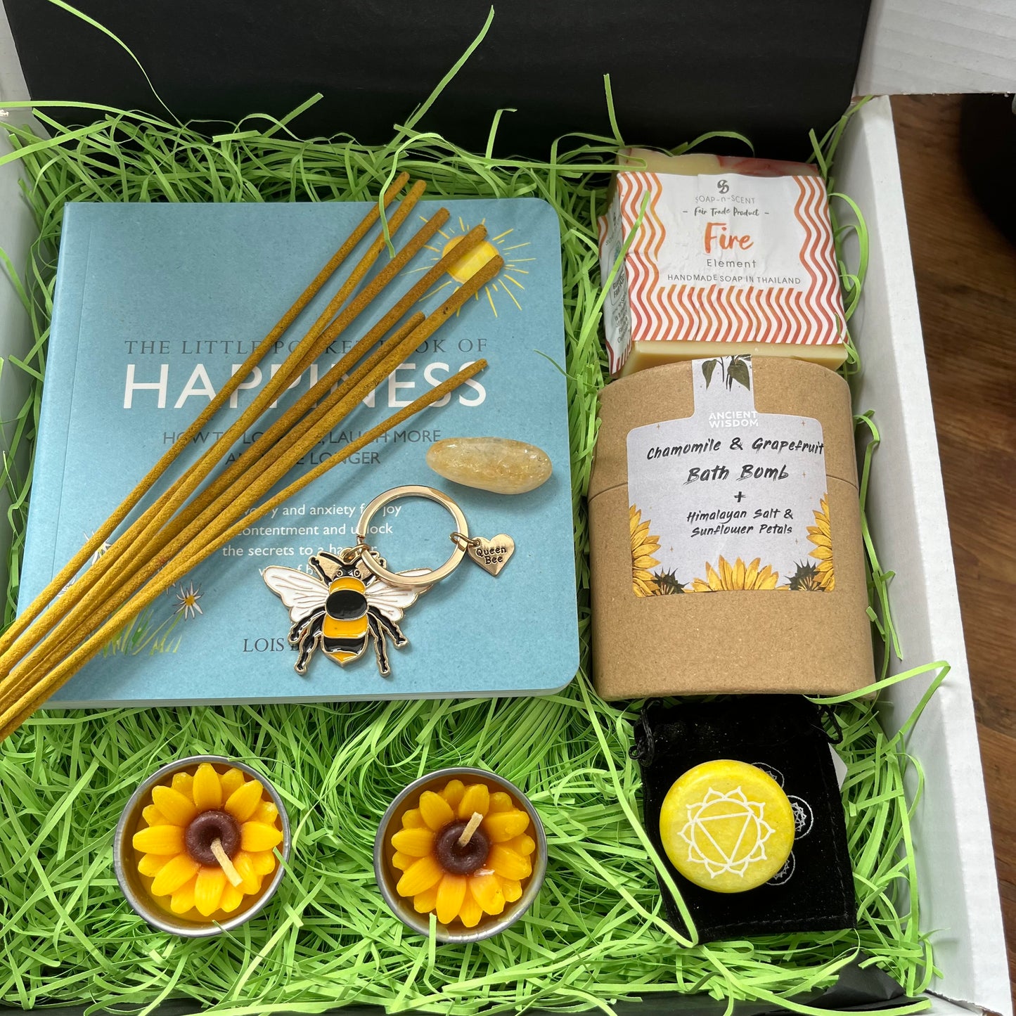 A curated gift box arrangement with a light blue book titled 'The Little Book of Happiness,' gold incense sticks, a bumblebee keychain, a wrapped bath bomb labeled 'Chamomile & Green Tea Bath Bomb,' two yellow flower-shaped tea lights, a yellow gemstone, and a black box with a white geometric emblem, all arranged in bright green paper filler.