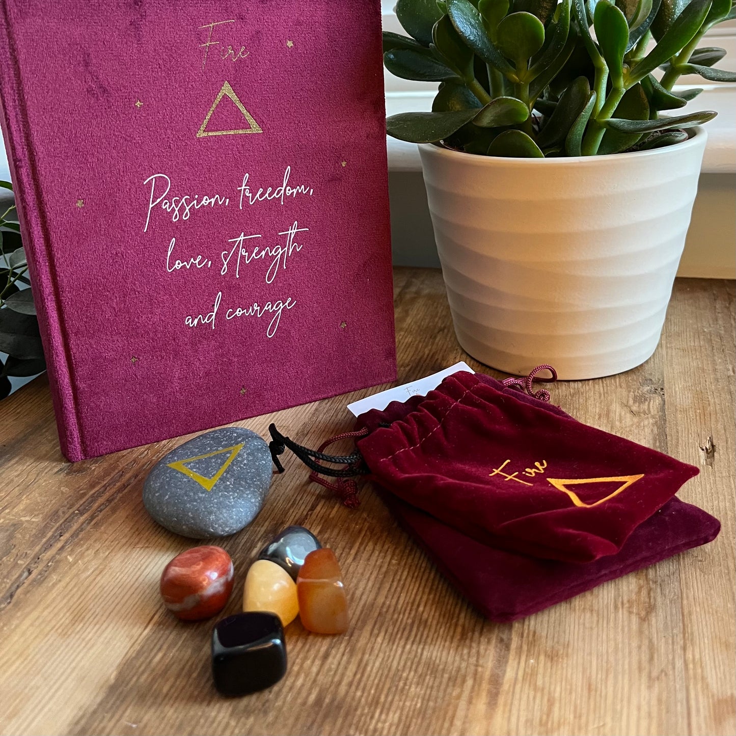 The contents of our 'Fire Element Gift Box' are shown on a wooden table. We can see a red velvet notebook with the word fire on the cover with the symbol for fire and inspirational words. A grey stone with the symbol for fire inscribed onto it. 5 tumblestones connected to the fire element Black Obsidian, yellow Aventurine, Carnelian, Hematite and red Jasper also shows 2 red velvet pouches one for the rune the other for the tumblestones.