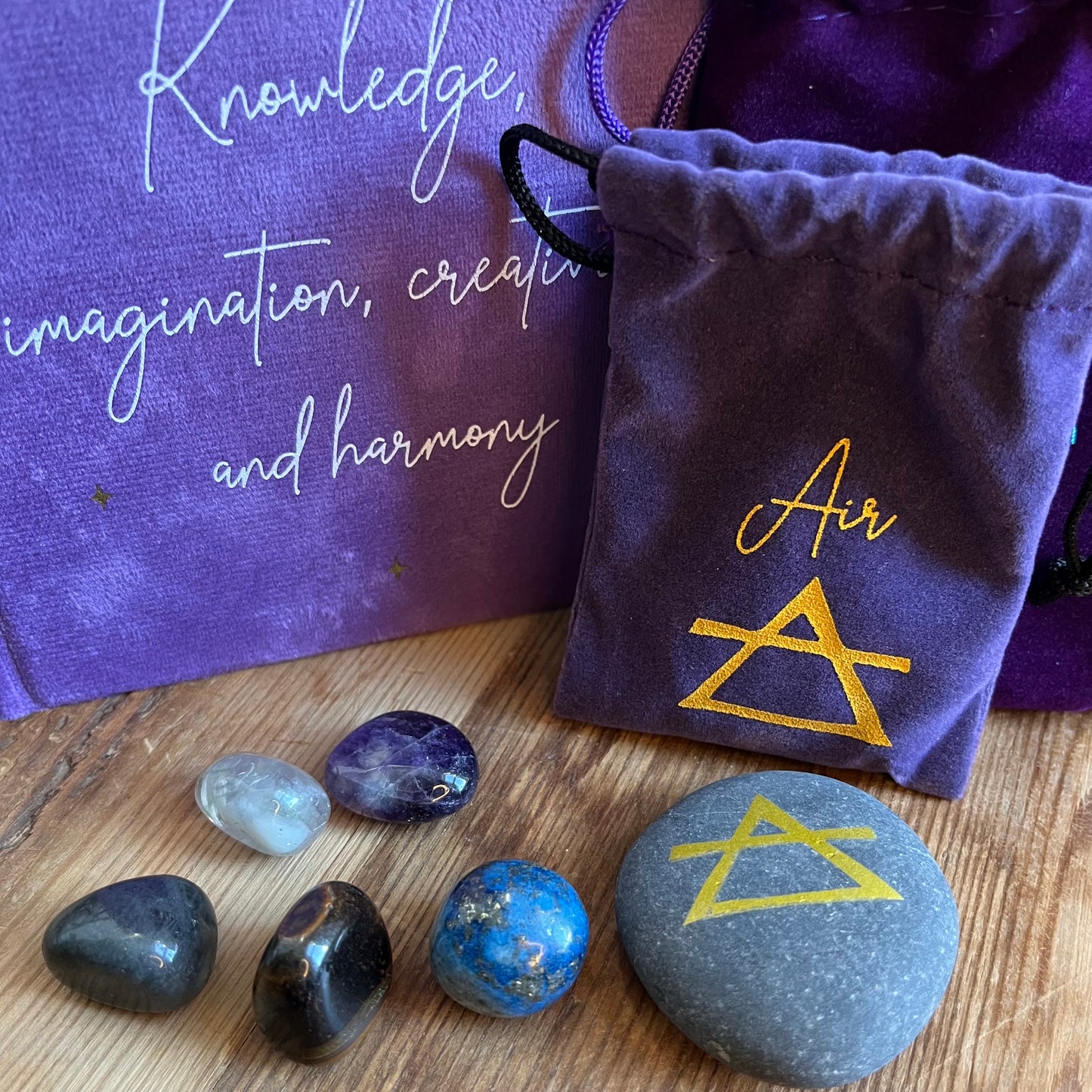 On a wooden table sits the contents of our 'Air Element Gift Box' a purple velvet notebook with inspiring words and the word air and the symbol for air. A grey rune stone decorated with the symbol for air alongside its purple velvet pouch also featuring the symbol for air. 5 tumble stones Lapis Lazuli, Amethyst, tigers eye, fluorite and labradorite, representing the element of air sit alongside their purple velvet pouch.