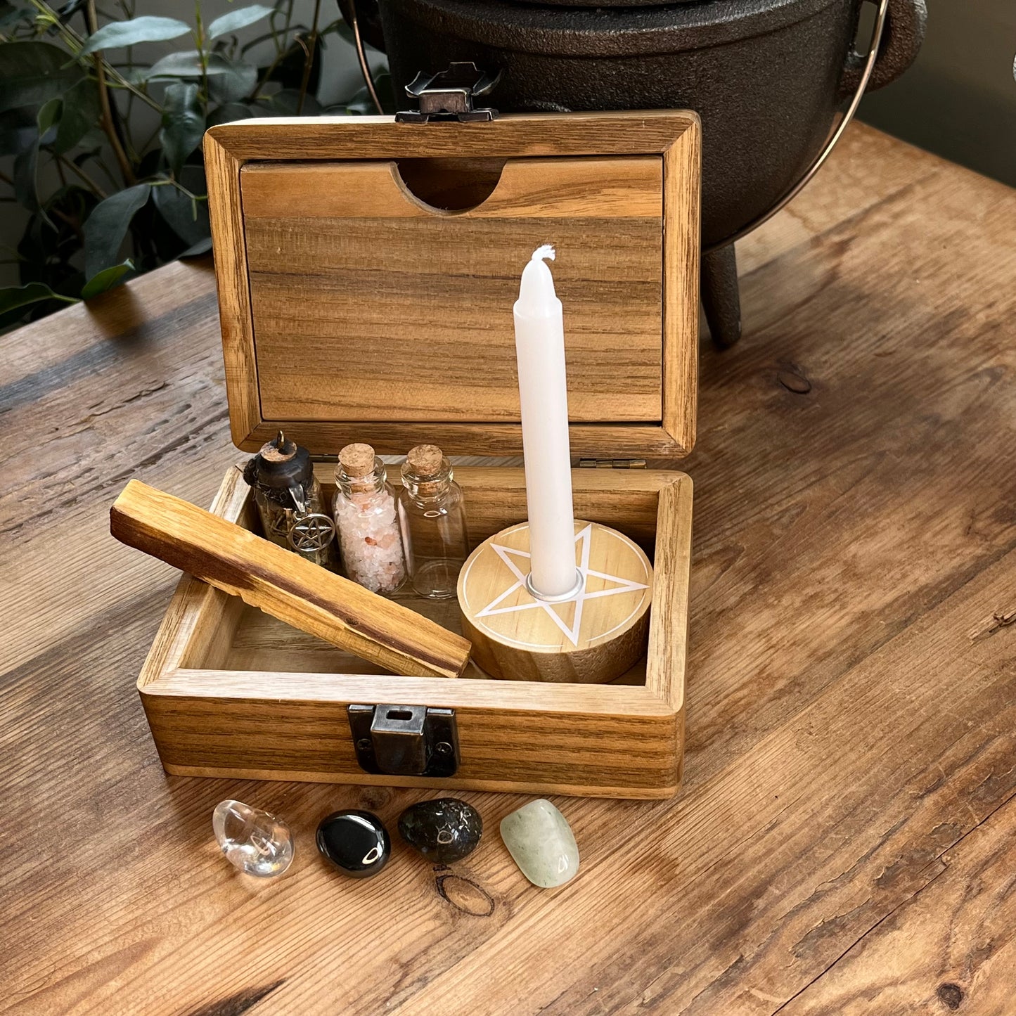 Inside the Wooden box with beautiful tree of life printed design on the lid, shows a separate magnetic compartment sat on a wooden table. The contents of the box can be seen. They are witchcraft or pagan tools. A white spell candle with a wooden candle holder with pentagram design. a palo santo smudge stick, 4 crystal tumble stones to represent the 4 elements, 3 spell bottles 1 containing herbs for protection, 1 containing salt for cleansing and 1 empty bottle.