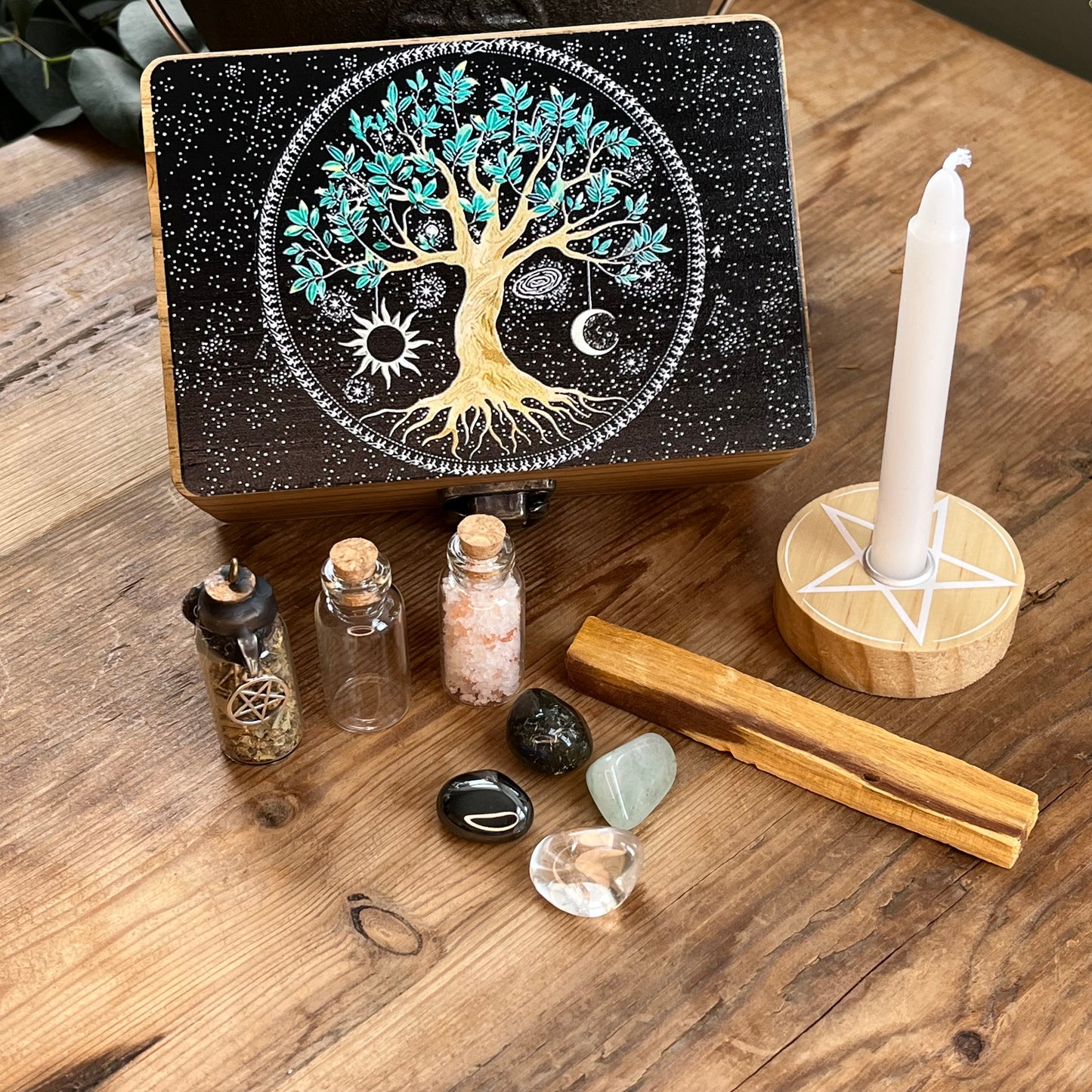 Wooden box with beautiful tree of life printed design on the lid on a table alongside the contents of the box, which are witchcraft or pagan tools. A white spell candle with a wooden candle holder with pentagram design. a palo santo smudge stick, 4 crystal tumble stones to represent the 4 elements, 3 spell bottles 1 containing herbs for protection, 1 containing salt for cleansing and 1 empty bottle.