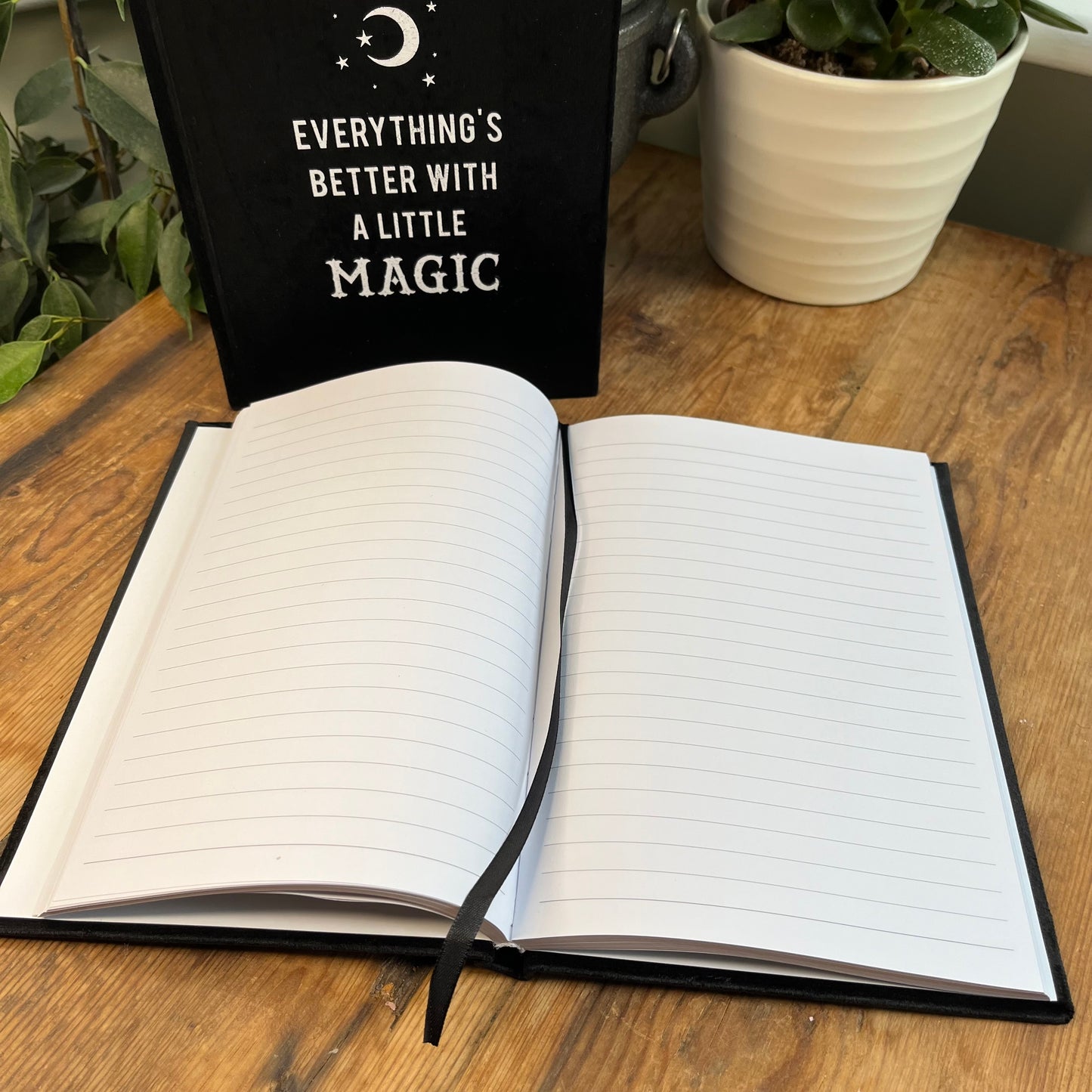 Black velvet notebook. Front cover has moon and stars design and the words 'everything's better with a little magic' The notebook is pictured with a second notebook that is open and the lined pages can be seen