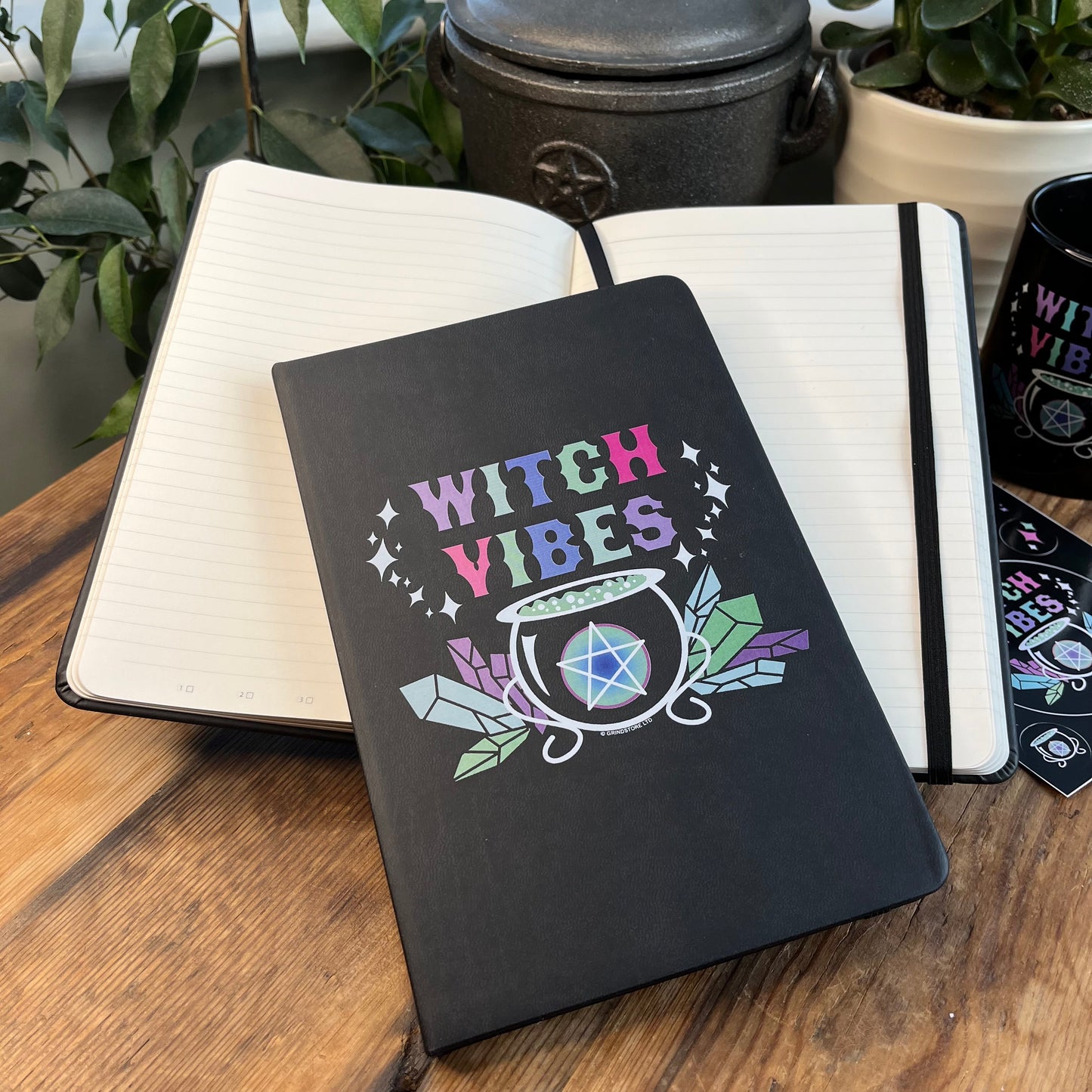 Black hard cover notebook. Front cover has a graphic of a cauldron, crystals and a pentagram and the words 'witch vibes ' in shades of purple, green, blue and pink. Image shows a closed notebook and an open notebook where the lined pages can be seen.