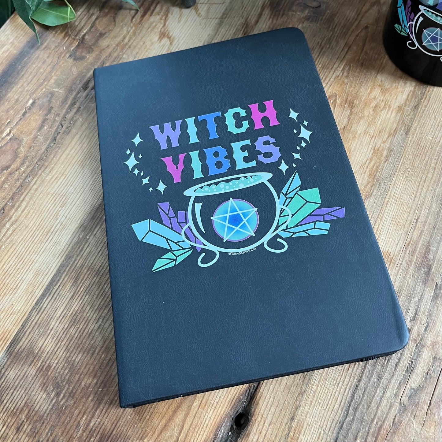 Black hard cover notebook. Front cover has a graphic of a cauldron, crystals and a pentagram and the words 'witch vibes ' in shades of purple, green, blue and pink.
