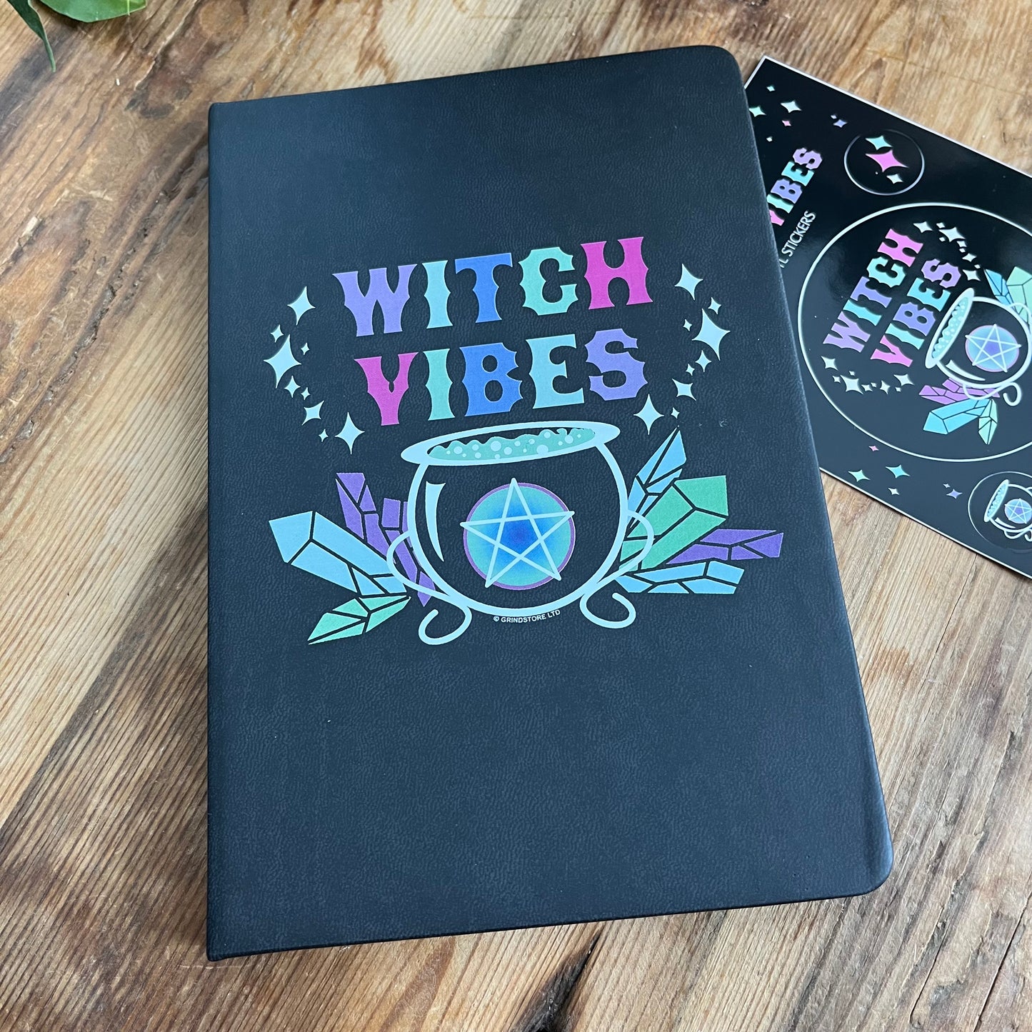 Black hard cover notebook. Front cover has a graphic of a cauldron, crystals and a pentagram and the words 'witch vibes ' in shades of purple, green, blue and pink.