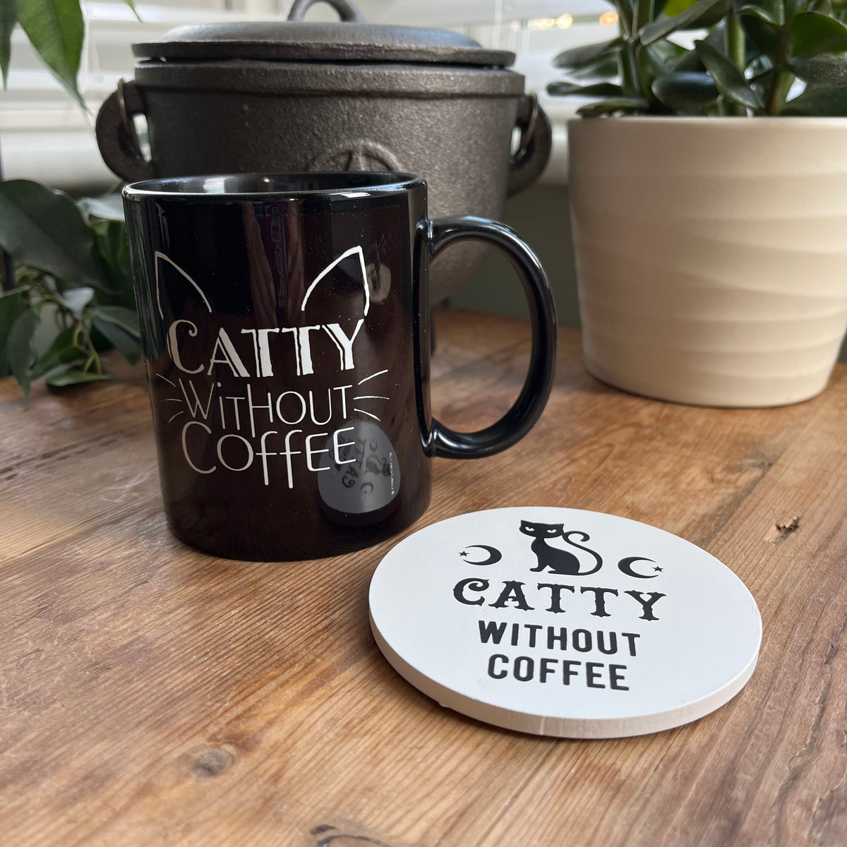 Black mug with the words 'catty without coffee' on a wooden table with a matchinh coaster