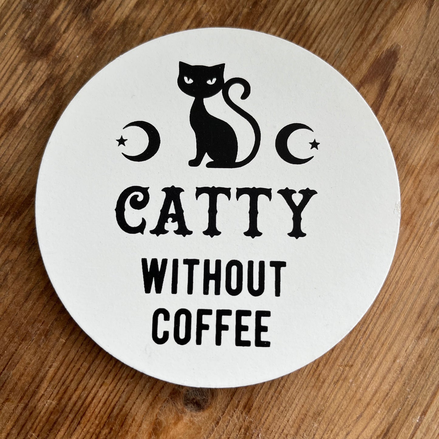 A round coaster with a black silhouette of a cat, the text 'CATTY Without Coffee', and decorative elements like stars and a crescent moon.