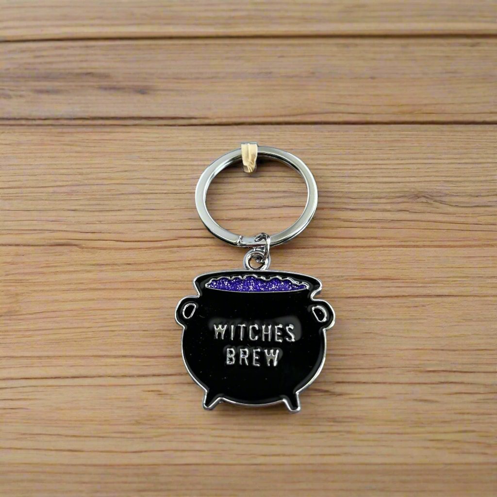 A Black cauldron with purple ligiud inside on a keyring. The words witches brew is written on the cauldron