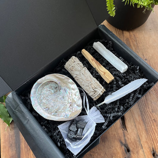 Black gift box exhibiting the contents of our 'sacred cleansing ritual gift box' - abalone shell, sage smoke stick, palo santo stick, rough raw selenite stick, 5 rough black tourmaline gemstones and a white goose feather. phtographed on a wooden table with a plant in the background