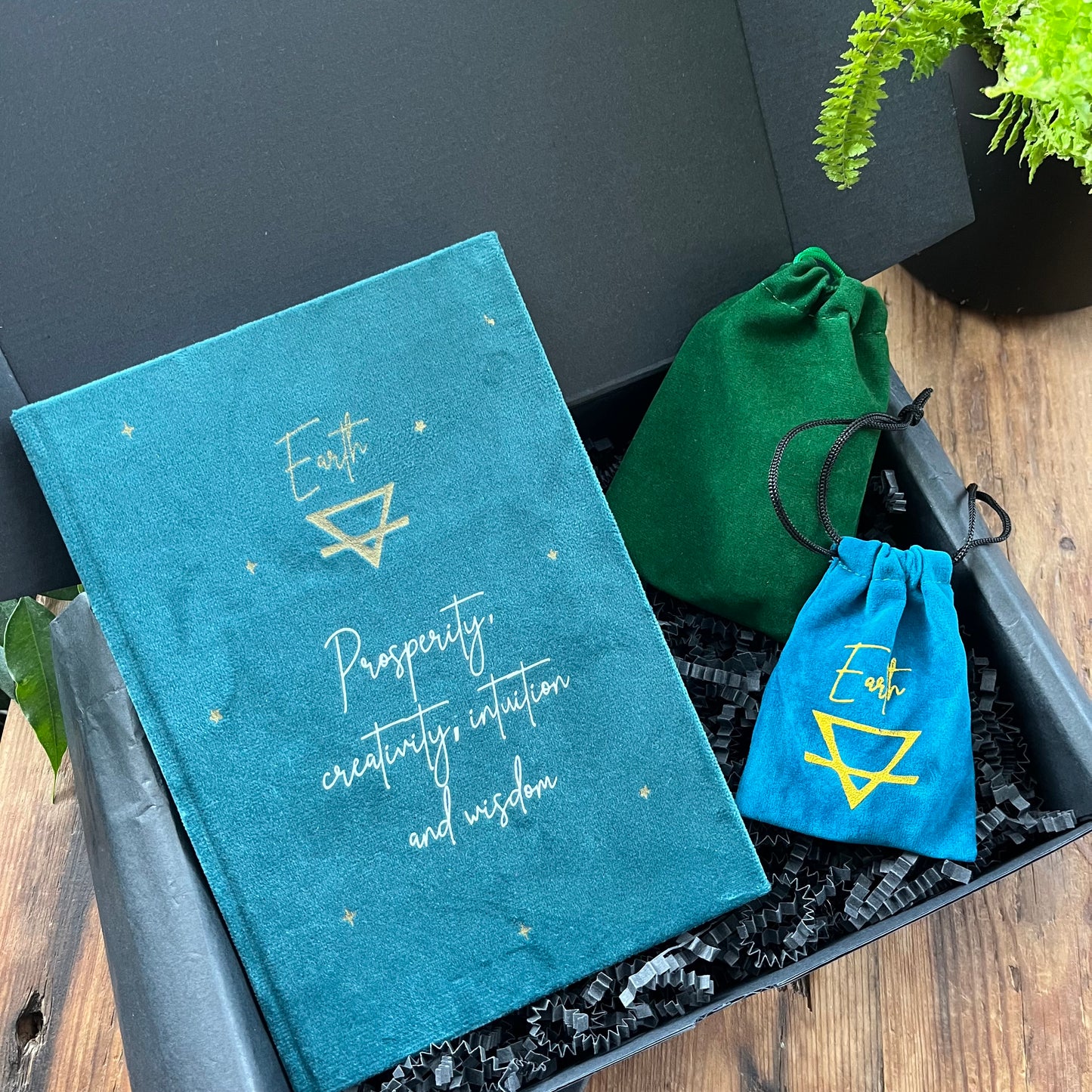 A Black gift box containing our 'Earth sign elemental gift box' - a Green velvet notebook featuring the elemental symbol for earth and inspiring words, two velvet pouches 1 containing a stone which features the elemental symbol for earth and the other contains tumbled stones associated with the element of earth