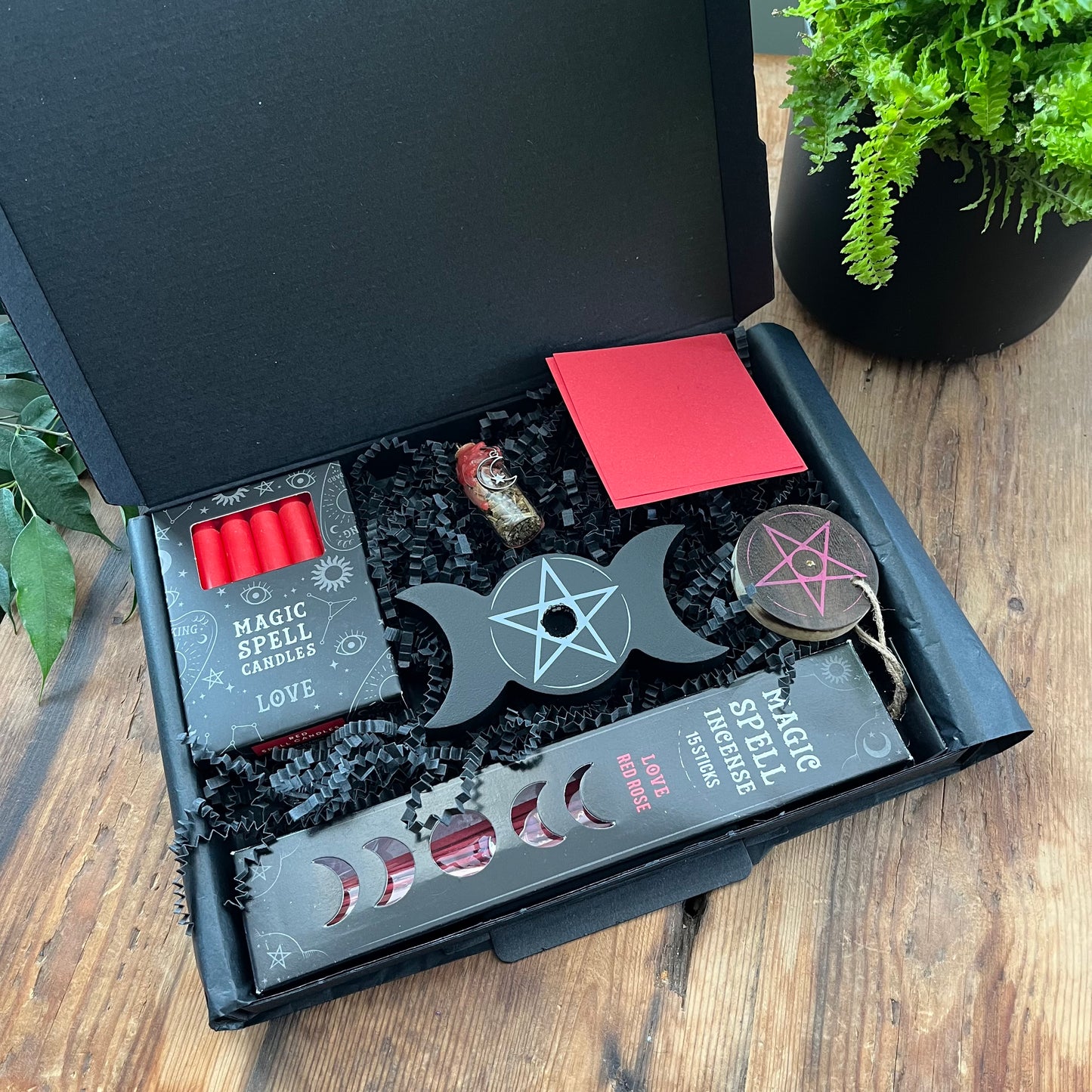 Black letterbox sized gifrt box containing our 'love spell kit''Love Spell Kit' - Pack of Love Incense sticks, and wooden pentagram incense holder, Pack of red magic spell candles, wooden black candle holder with pentagram design, squares of red paper and a "Love' spell bottle containing herbs and sealed with red wax and a charm. Photographed on a wooden table with a plant in the background