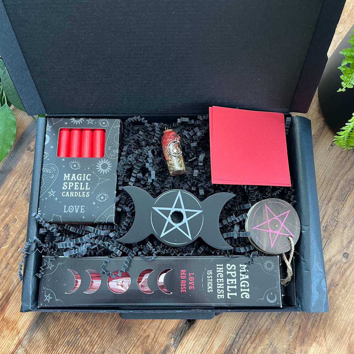 Black letterbox sized gifrt box containing our 'love spell kit''Love Spell Kit' - Pack of Love Incense sticks, and wooden pentagram incense holder, Pack of red magic spell candles, wooden black candle holder with pentagram design, squares of red paper and a "Love' spell bottle containing herbs and sealed with red wax and a charm. Photographed on a wooden table with a plant in the background