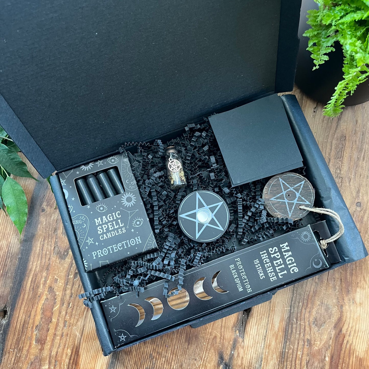 Black letterbox size gift box containing 'protection spell kit''Protection Spell Kit' Pack of black opium scented incense sticks for protection and wooden pentagram incense holder, Pack of black magic spell candles, wooden black candle holder, in triple moon and pentagram design. squares of black paper and a "protection" spell bottle containing herbs and sealed with black wax and a charm. Photographed on a wooden table with a plant in the background