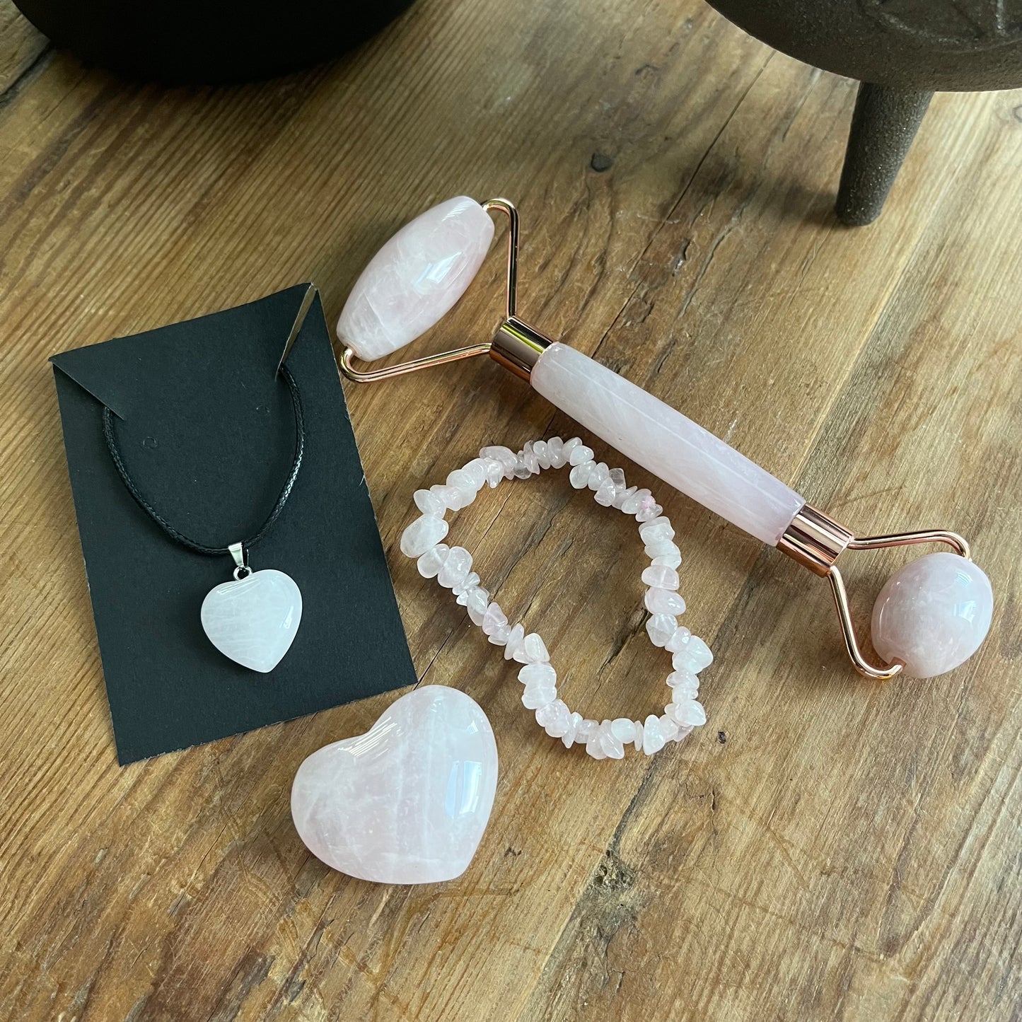 contents of the Rose Quartz selfcare gift box sat on a wooden table.  rose quartz face roller, rose quartz chip bracelet, a rose quartz heart , and a rose quartz pendent heart necklace. Plus information cards with the properties of rose quartz and benefits of a face roller.