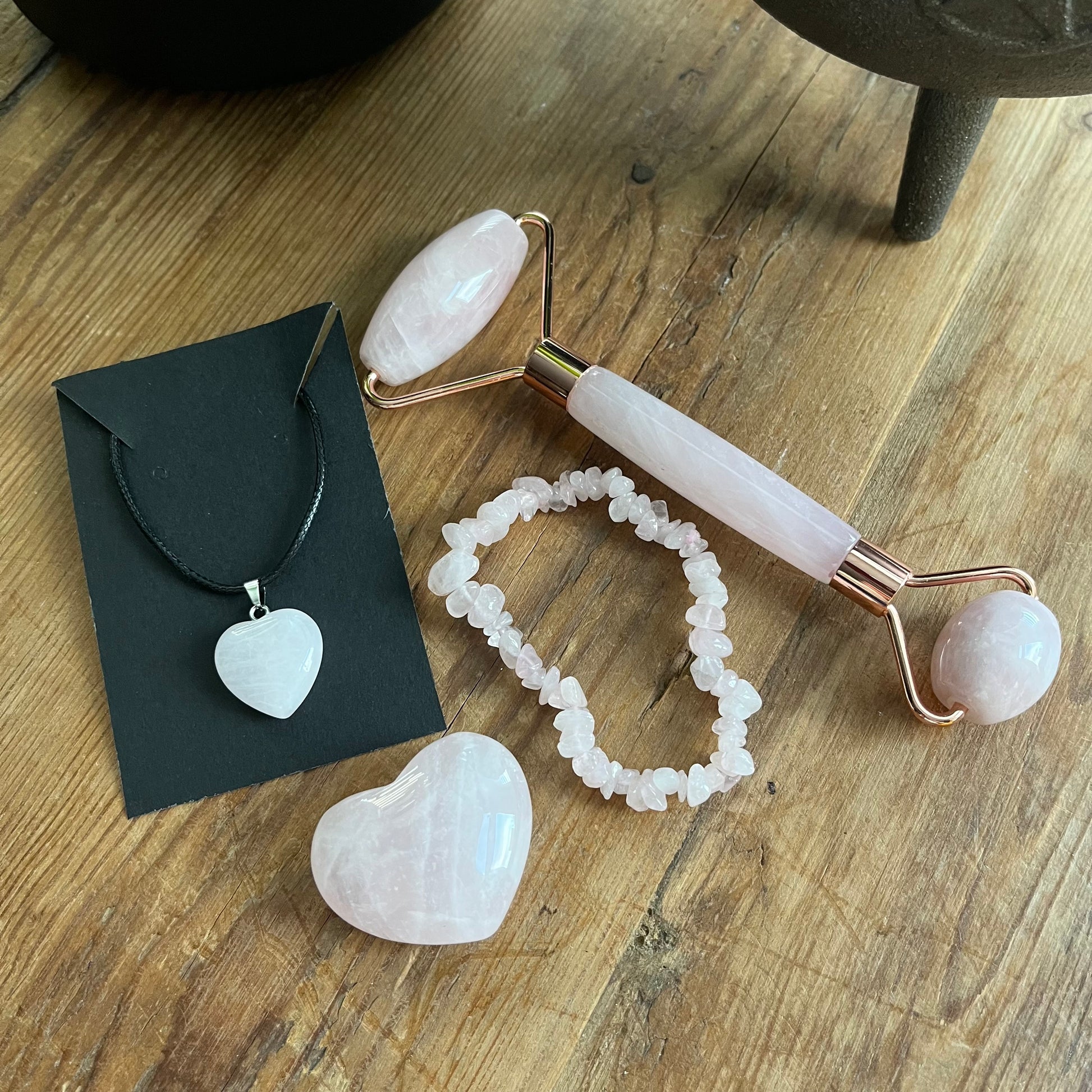 contents of the Rose Quartz selfcare gift box sat on a wooden table.  rose quartz face roller, rose quartz chip bracelet, a rose quartz heart , and a rose quartz pendent heart necklace. Plus information cards with the properties of rose quartz and benefits of a face roller.