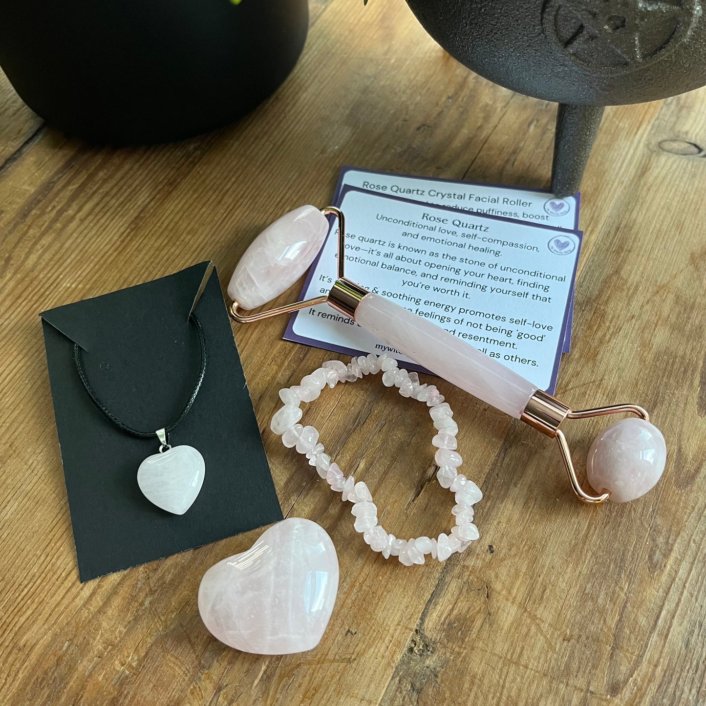 contents of the Rose Quartz selfcare gift box sat on a wooden table.  rose quartz face roller, rose quartz chip bracelet, a rose quartz heart , and a rose quartz pendent heart necklace. Plus information cards with the properties of rose quartz and benefits of a face roller.