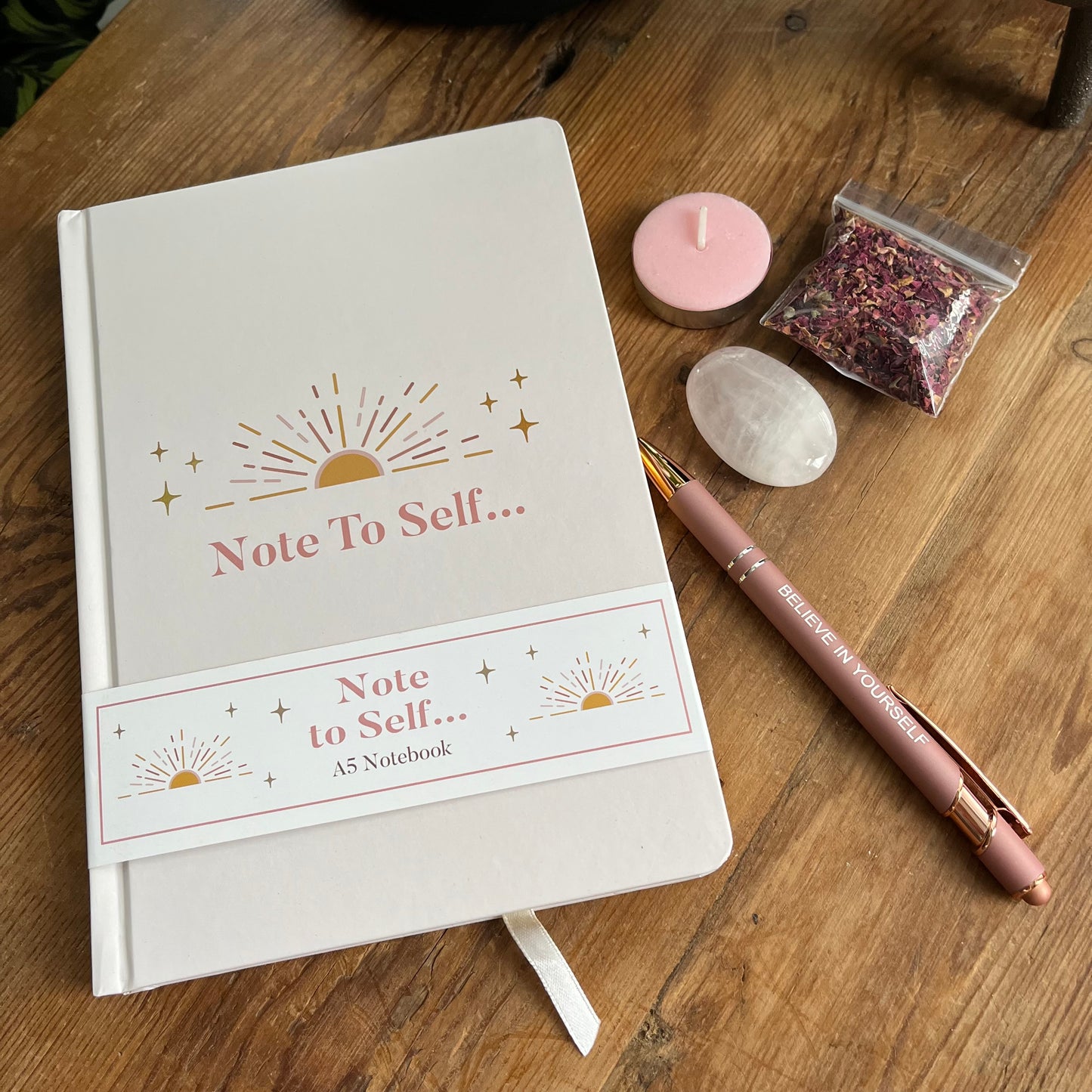 Contents of Note to self - ritual for self -love gift box. Pale pink & gold A5 notebook with the words note to self, a rose gold coloured pen with a motivational message written on the side, a rose quartz palm stone, a pink tealight candle and a packet of red rose petals.