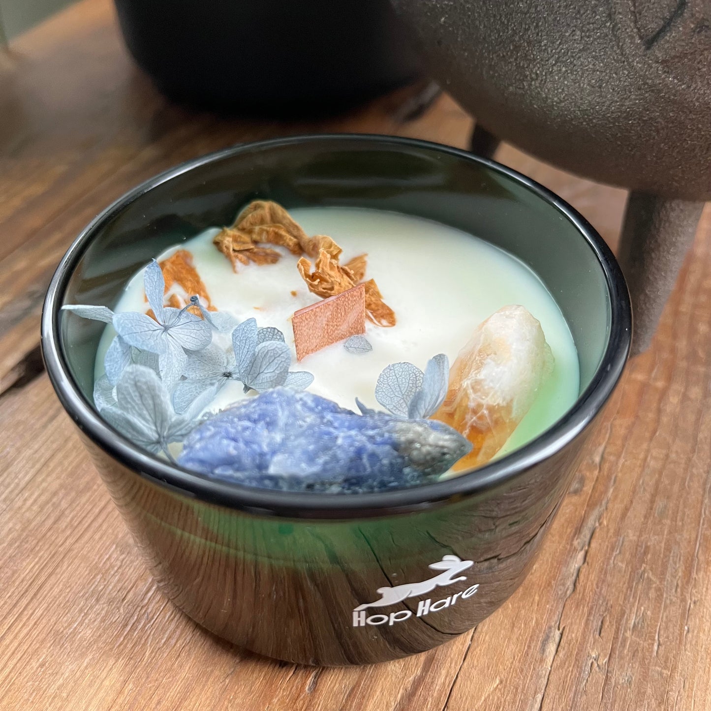 Hop Hare Sun Candle with wooden wick, blue flowers and arge citrine and sodalite crystals. Candle itself is white and set in a dark green glass jar.