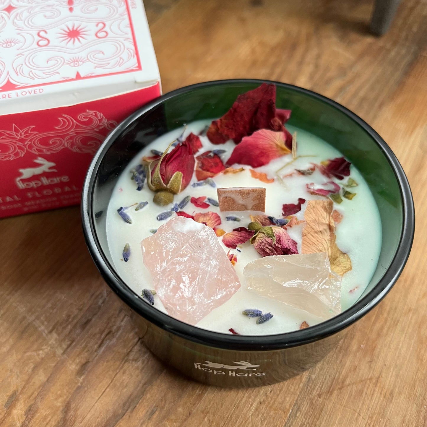 Top of the hop hare Lovers candle shows a wooden wick, large rose quartz crystals, red rose bud and rose petals.