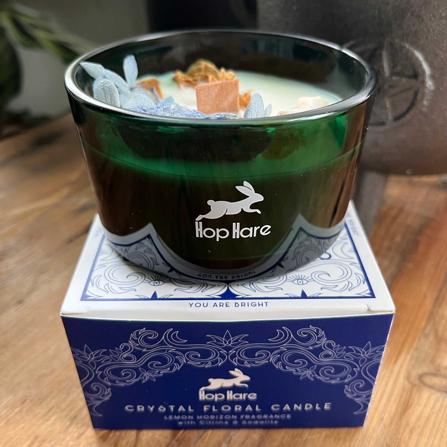 Hop Hare Sun Candle with wooden wick, blue flowers and large citrine and sodalite crystals. Candle itself is white and set in a dark green glass jar. The Candle is shown atop of it's blue and white decorative cardboard presentation box.