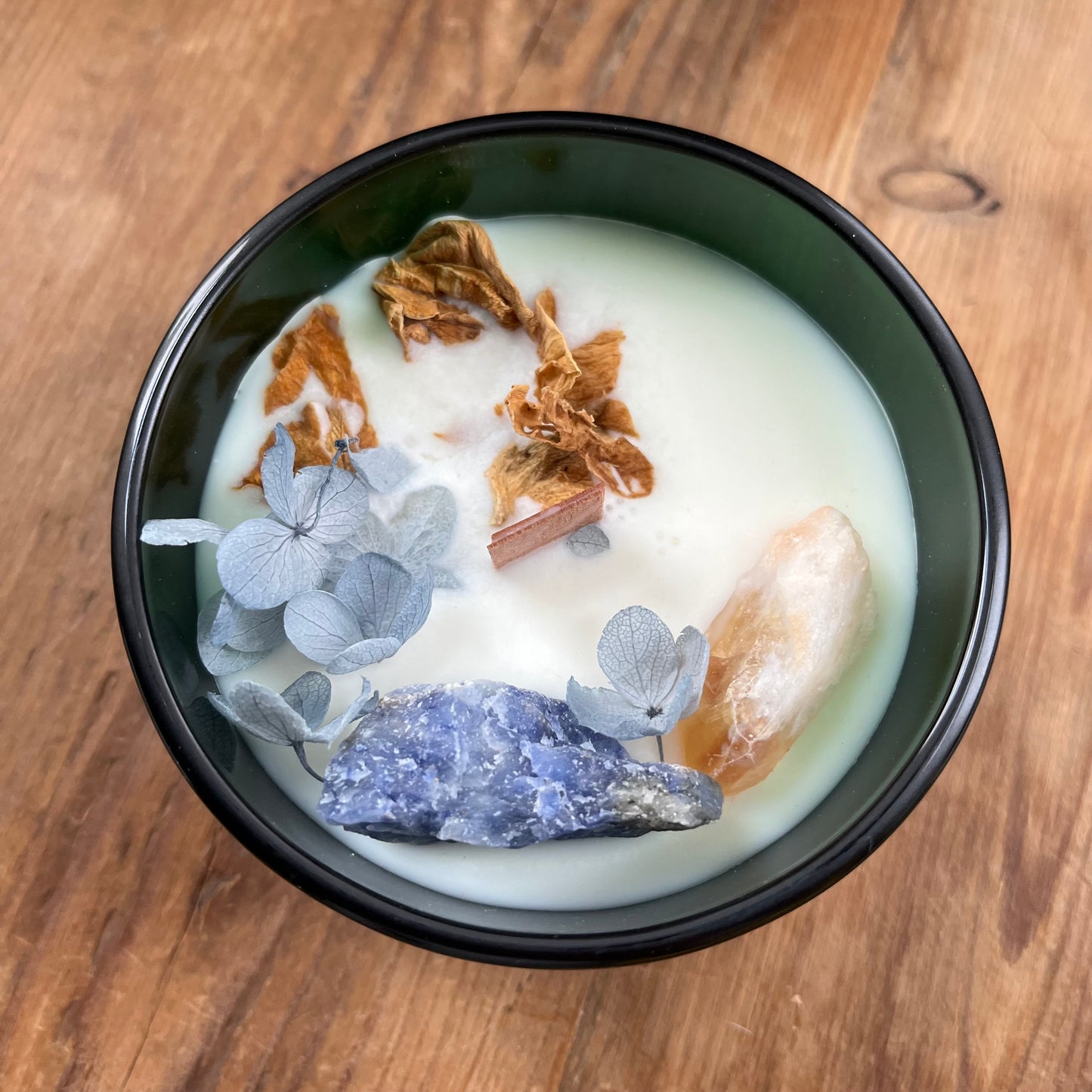 Hop Hare Sun Candle with wooden wick, blue flowers and large citrine and sodalite crystals. Candle itself is white and set in a dark green glass jar.