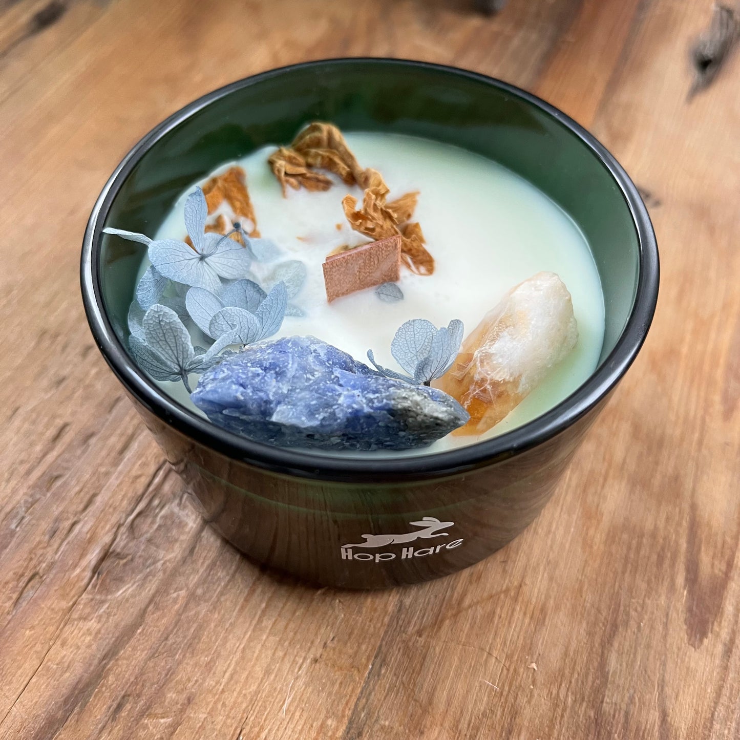 Hop Hare Sun Candle with wooden wick, blue flowers and large citrine and sodalite crystals. Candle itself is white and set in a dark green glass jar.