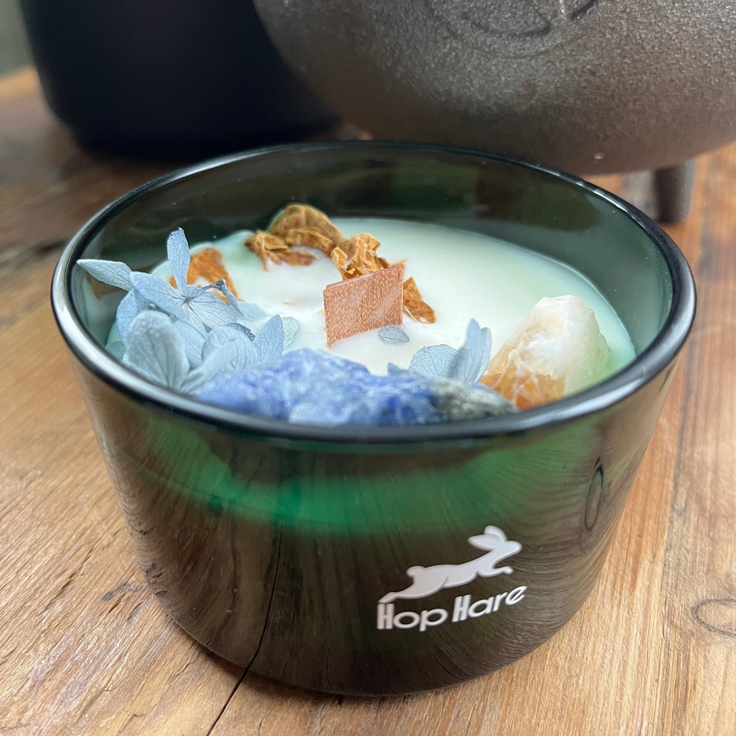 Hop Hare Sun Candle with wooden wick, blue flowers and large citrine and sodalite crystals. Candle itself is white and set in a dark green glass jar.