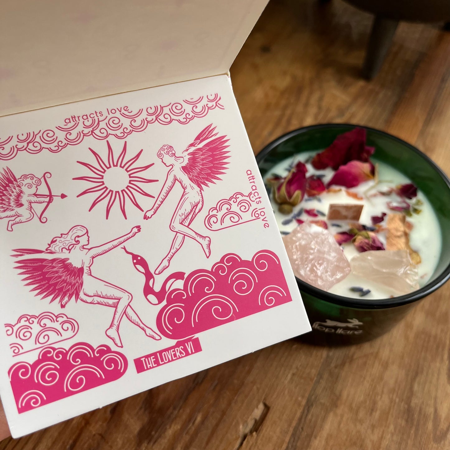 Top of the hop hare Lovers candle shows a wooden wick, large rose quartz crystals, red rose bud and rose petals. The candle itself is white and set in a dark green glass Jar. The candle is shown next to the accompanying tarot card.