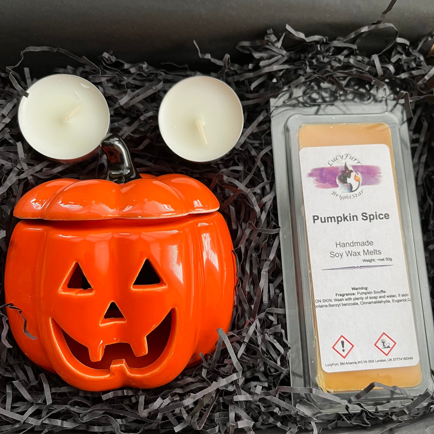 Pumpkin spice wax melt packaging with a jack-o'-lantern-shaped container on black shredded paper.