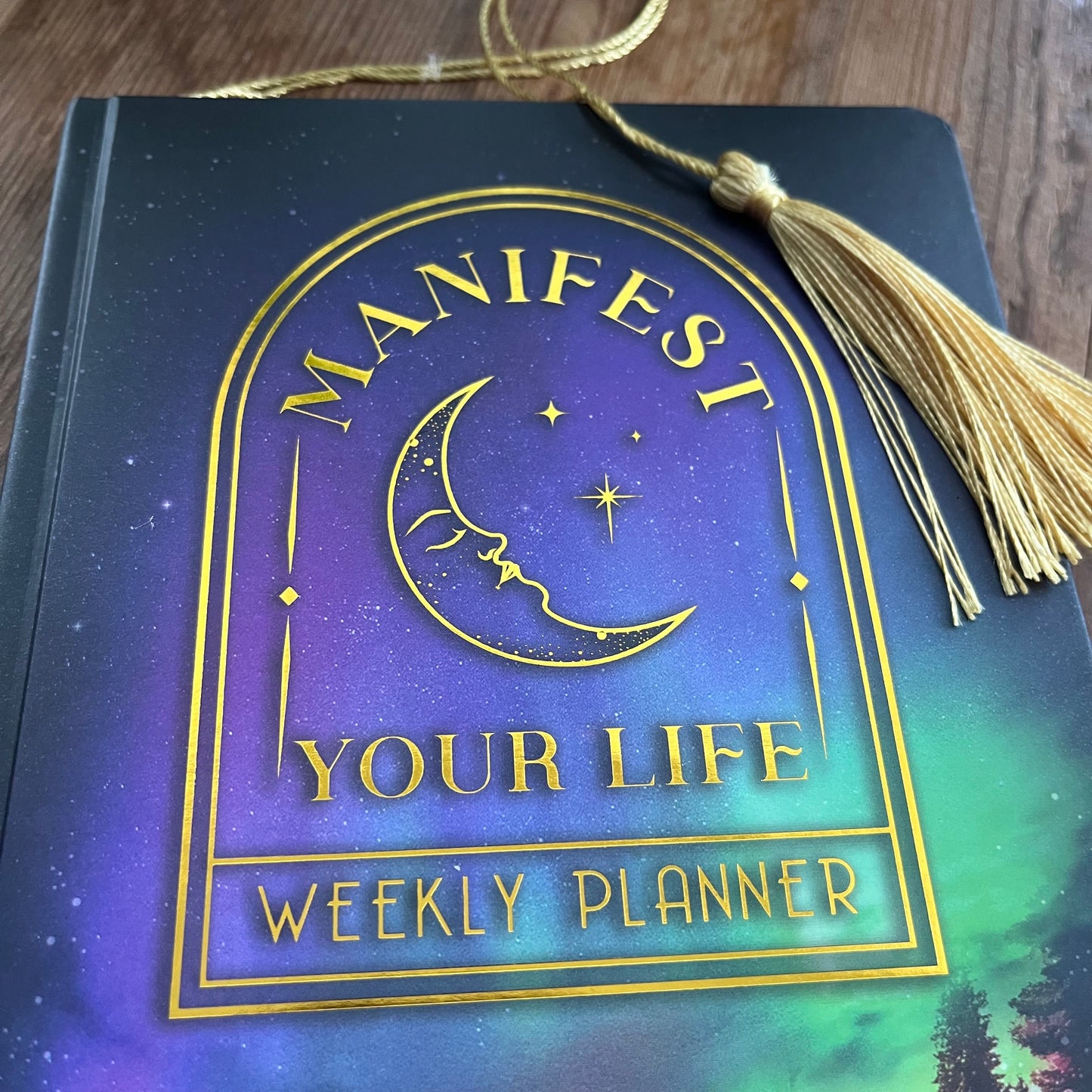 Weekly planner book titled 'Manifest Your Life' with a tassel on a wooden surface