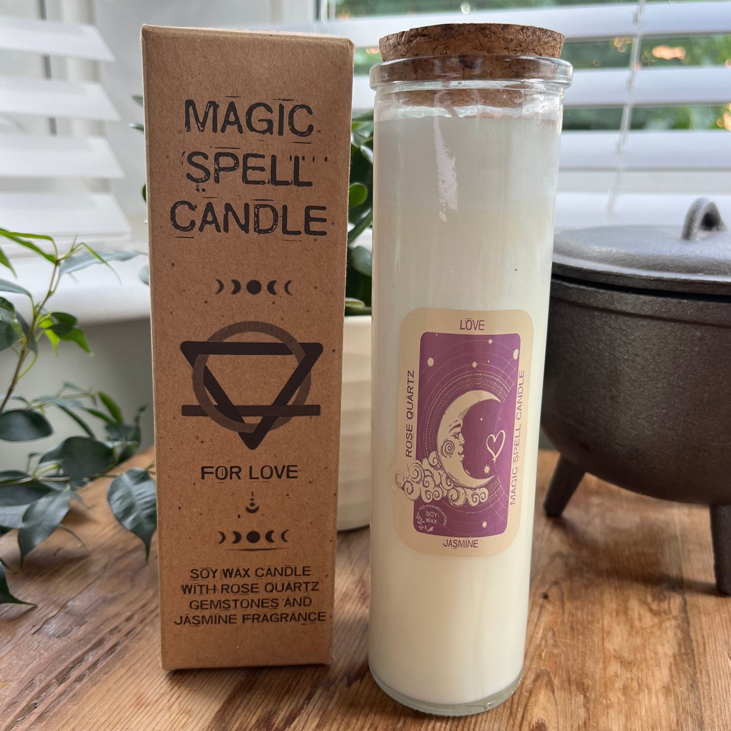 Tall glass spell candle with cork lid. The label on the candle features a crescent moon and love heart and the words Love, rose quartz and Jasmine. The candle itself is white with rose quartz gemstones embedded within it. Alongside the glass Magic spell candle is it's brown cardboard box which has the words magic spell candle for love, soy wax candle with rose quartz gemstones and Jasmine fragrance.