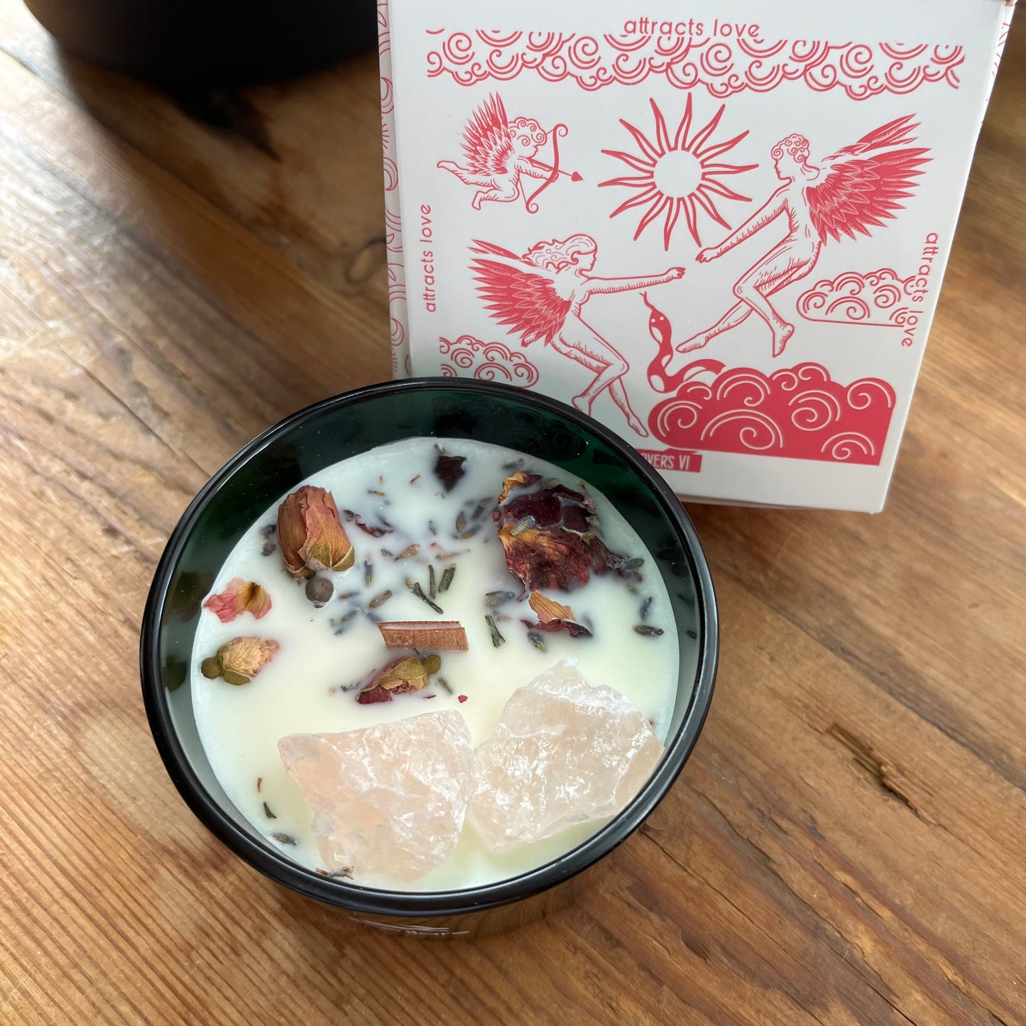 Candle in a black container with decorative elements on a wooden surface, next to a card with angel illustrations.