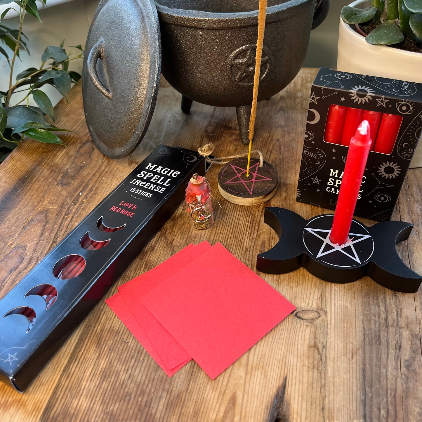 our 'love spell kit''Love Spell Kit' - Pack of Love Incense sticks, and wooden pentagram incense holder, Pack of red magic spell candles, wooden black candle holder with pentagram design, squares of red paper and a "Love' spell bottle containing herbs and sealed with red wax and a charm. Photographed on a wooden table with a plant in the background
