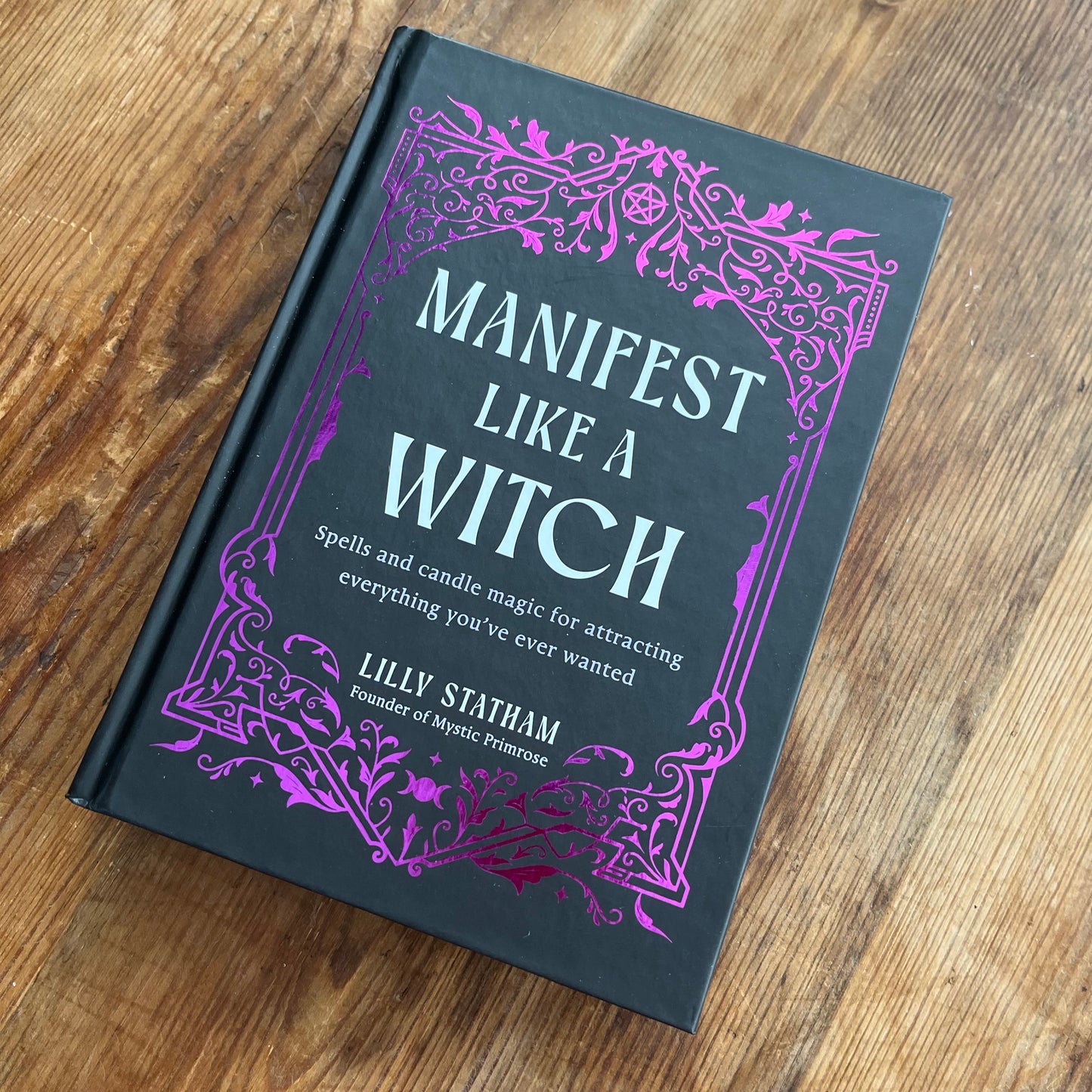 A hardcover book titled 'Manifest Like a Witch' by Lilly Statham, featuring a black cover with ornate purple and white gothic-style text and border designs, photographed on a wooden tabletop.