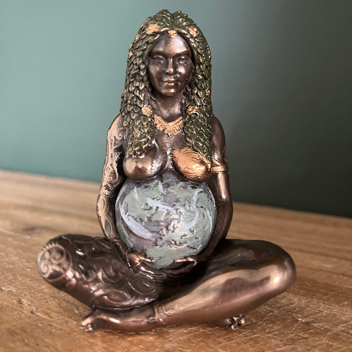 A bronze-finished figurine of a pregnant woman seated cross-legged, holding a blue and white marbled globe. The sculpture features warm metallic bronze tones with textured gold highlights on the hair, chest, and arms, set against a dark green wall and a wooden tabletop.