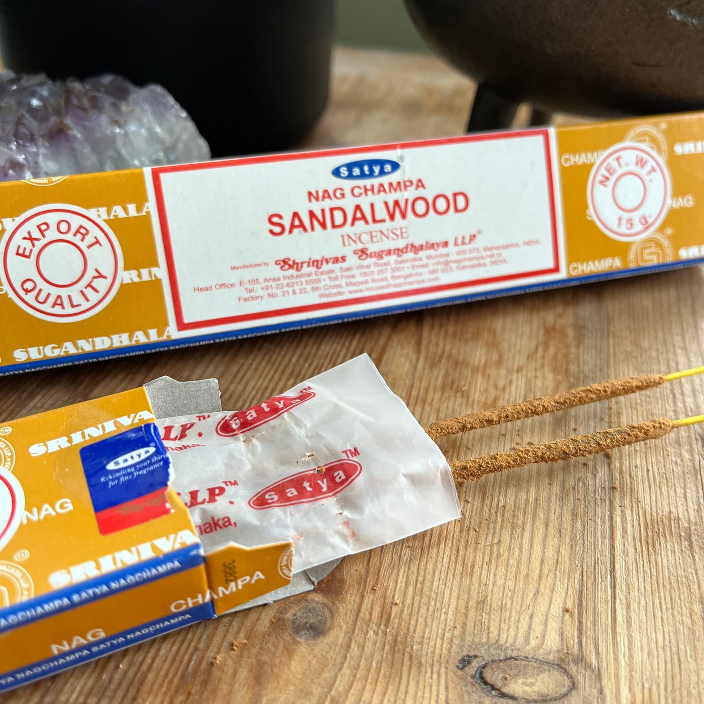 2 boxes of Satya Nag Champa Sandalwood incense, with branding and product information displayed on the packaging, placed on a wooden surface. One box is open and the incense sticks are peeking out