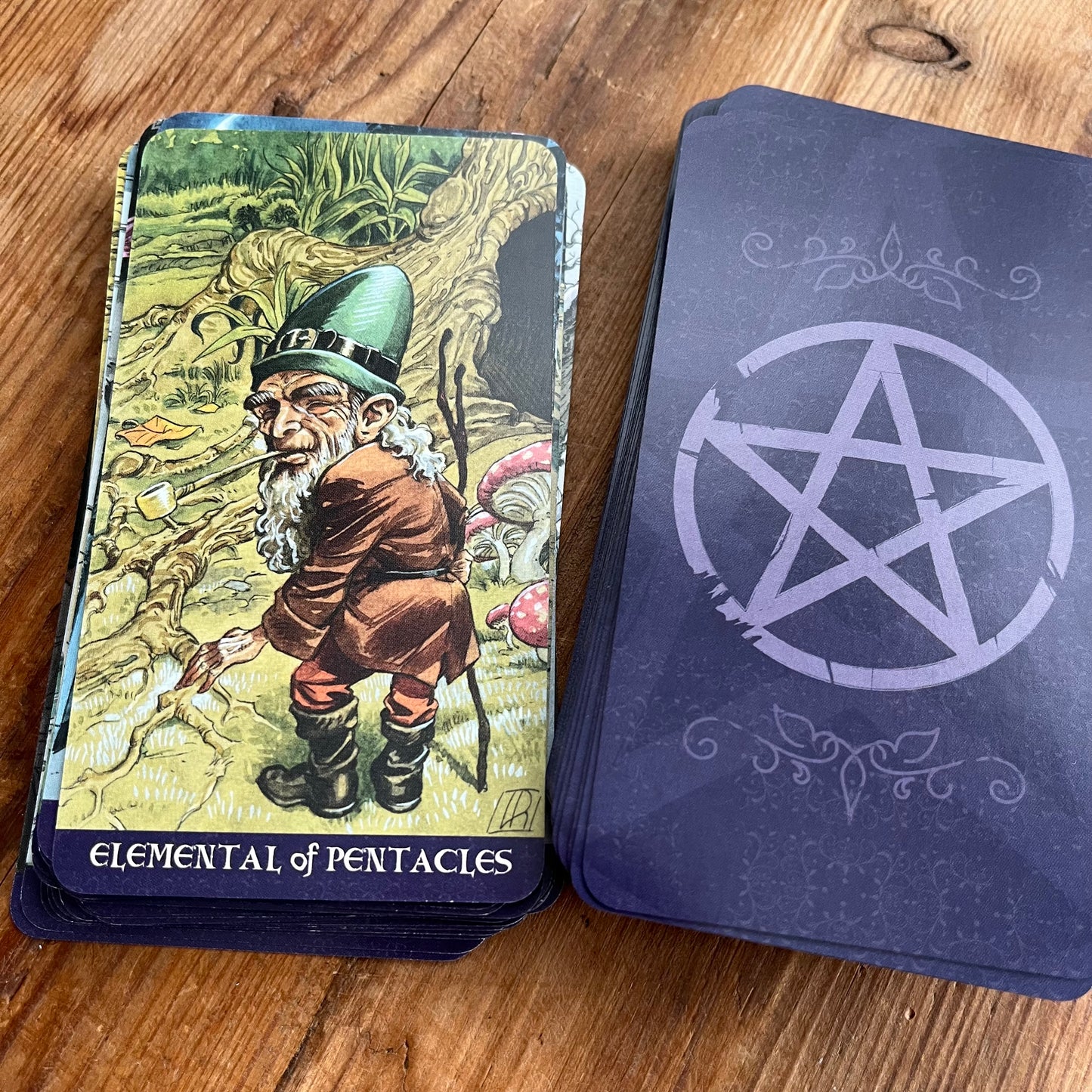 A deck of tarot cards labeled 'Elemental of Pentacles' with a colorful illustrated front and a dark blue back featuring a large pentagram symbol, placed on a wooden surface.