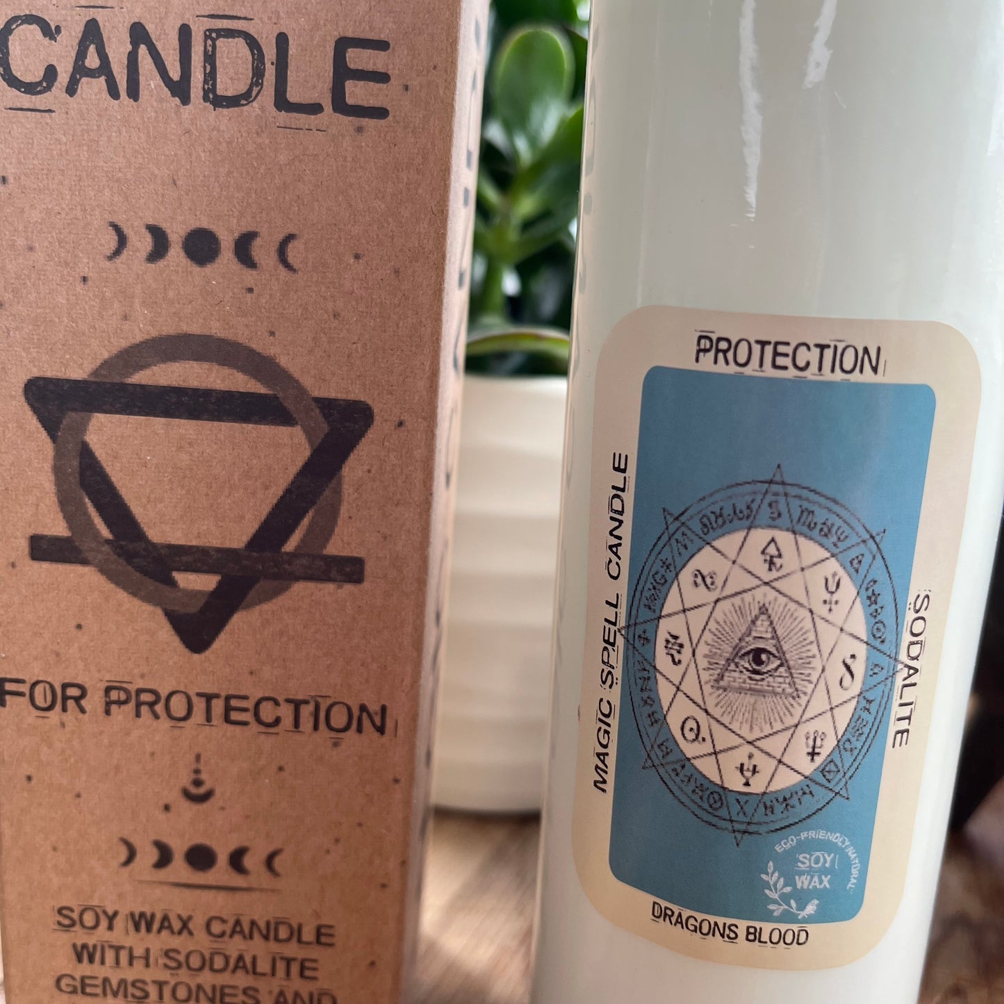 Tall glass spell candle with cork lid. The label on the candle features an image of the all seeing eye and the words Protection Sodalite and Dragons Blood. The candle itself is white with Sodalite gemstones embedded within it. Alongside the glass Magic spell candle is it's brown cardboard box which has the words magic spell candle for protection, soy wax candle with Sodalite gemstones and Dragons Blood fragrance.