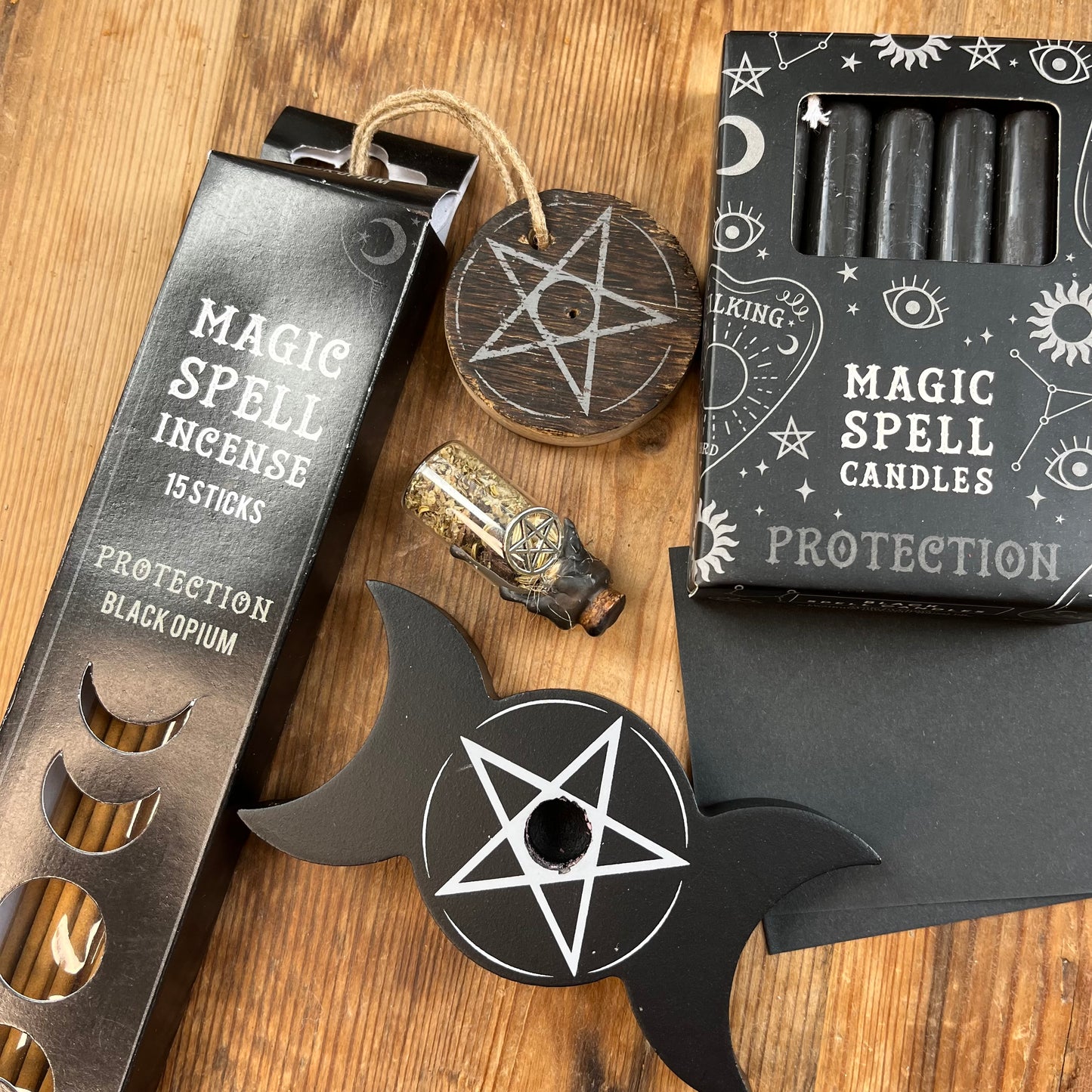 'Protection Spell Kit' Contents displayed on a wooden table. Pack of black opium scented incense sticks for protection and wooden pentagram incense holder, Pack of black magic spell candles, wooden black candle holder, in triple moon and pentagram design. squares of black paper and a "protection" spell bottle containing herbs and sealed with black wax and a charm.