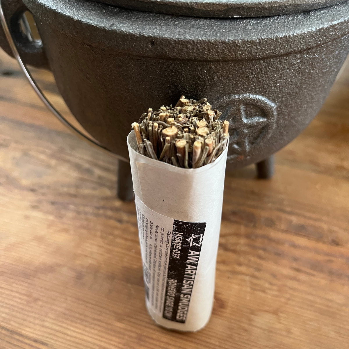 A bundle of dried herbs or spices wrapped in a white paper sleeve with a black label, placed in front of a dark cast iron pot on a wooden surface.