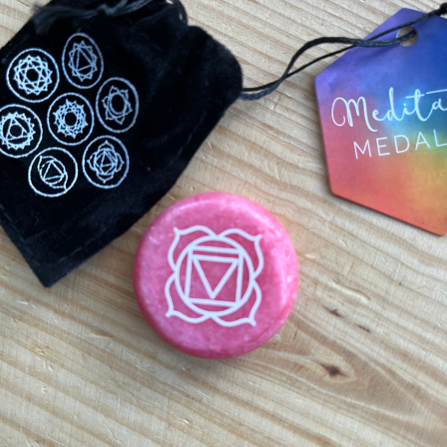 A red circular flat meditation stone with the root chakra symbol on it, with a black velvet pouch which has the 7 chakra smbols on it
