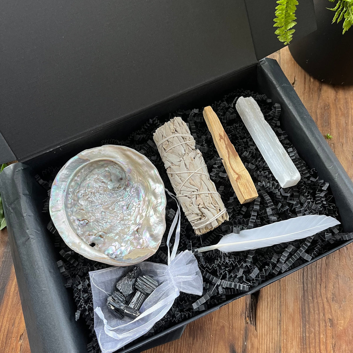 A black gift box exhibiting the contents of our 'sacred cleansing ritual gift box' - abalone shell, sage smoke stick, palo santo stick, rough raw selenite stick, 5 rough black tourmaline gemstones and a white goose feather. Photographed on a wooden table with a plant in the background