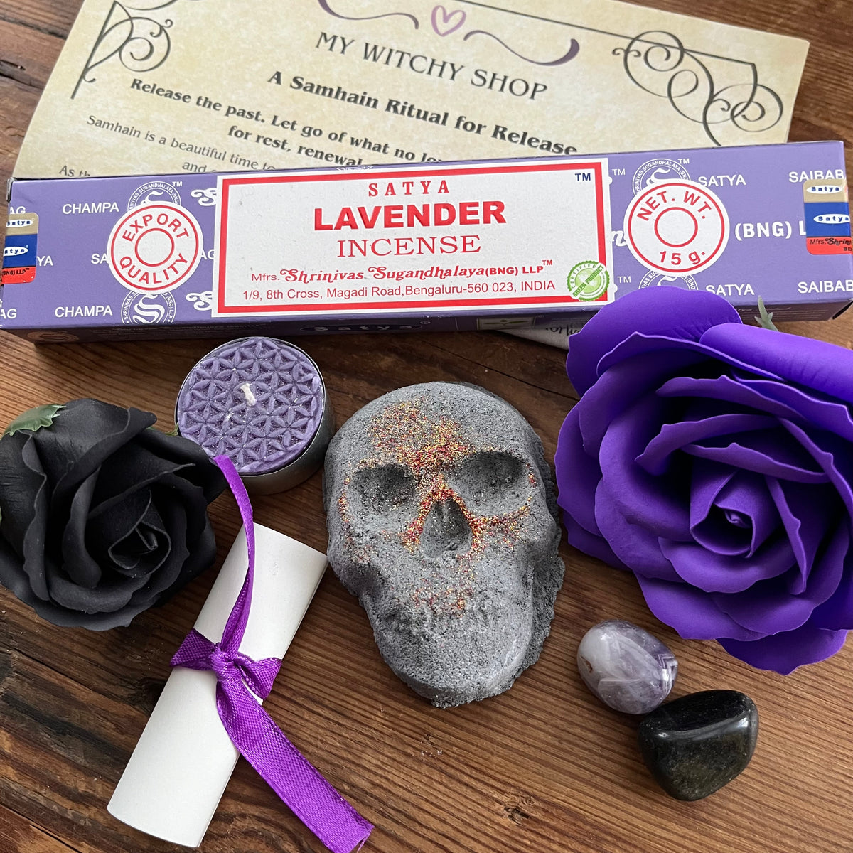 lavender incense sticks, package tied with a purple ribbon, a small white scroll, a skull shaped bath bomb, a purple rose and black rose craft soap, a purple candle and a purple organza bag containing crystals . An information sheet reading 'MY WITCHY SHOP' is placed beside the contents on a wooden surface.